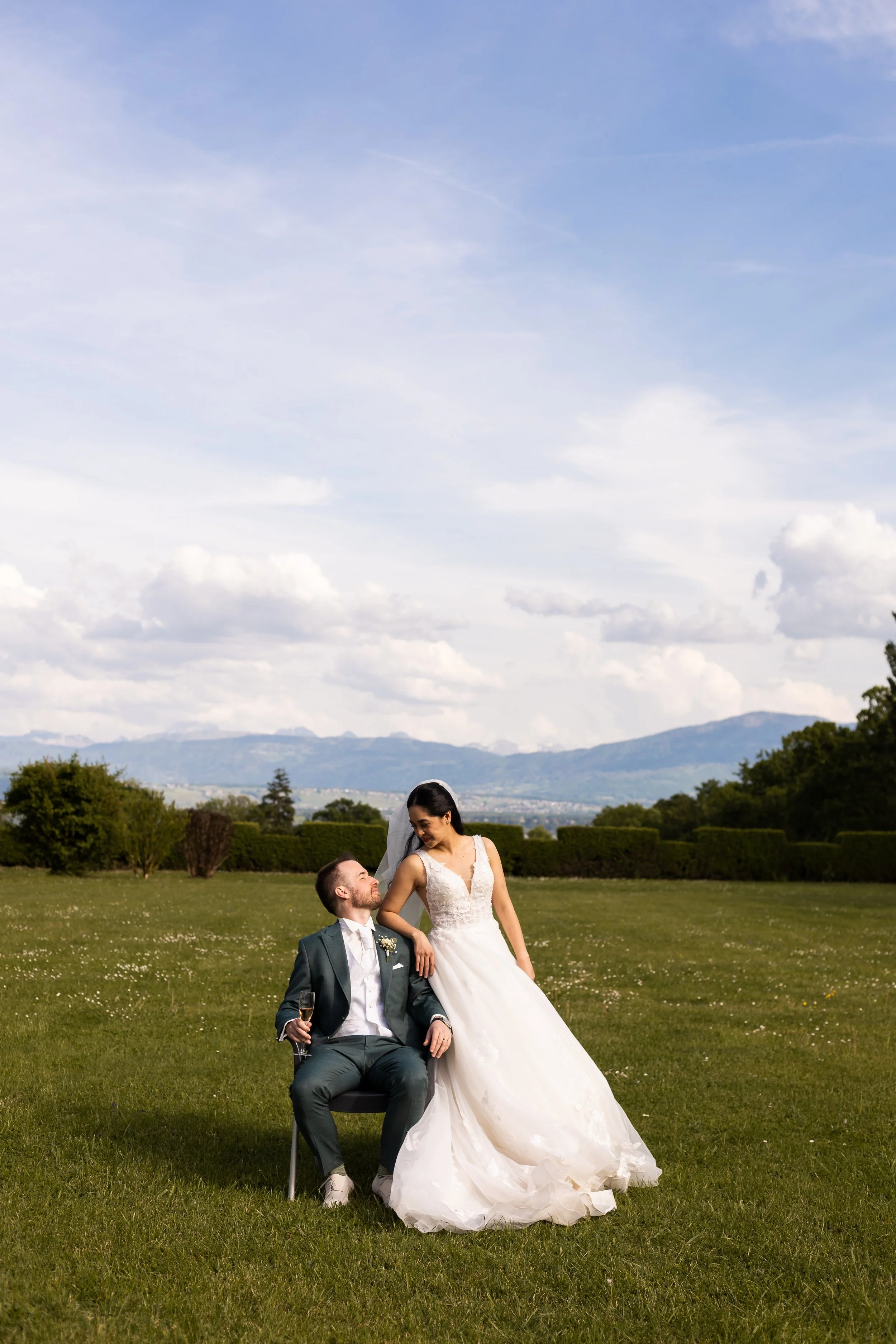 Un couple de mariés dans un champ verdoyant avec un paysage de montagnes au loin, la mariée debout à côté du marié qui est assis, ils partagent un moment heureux lors de leur mariage.