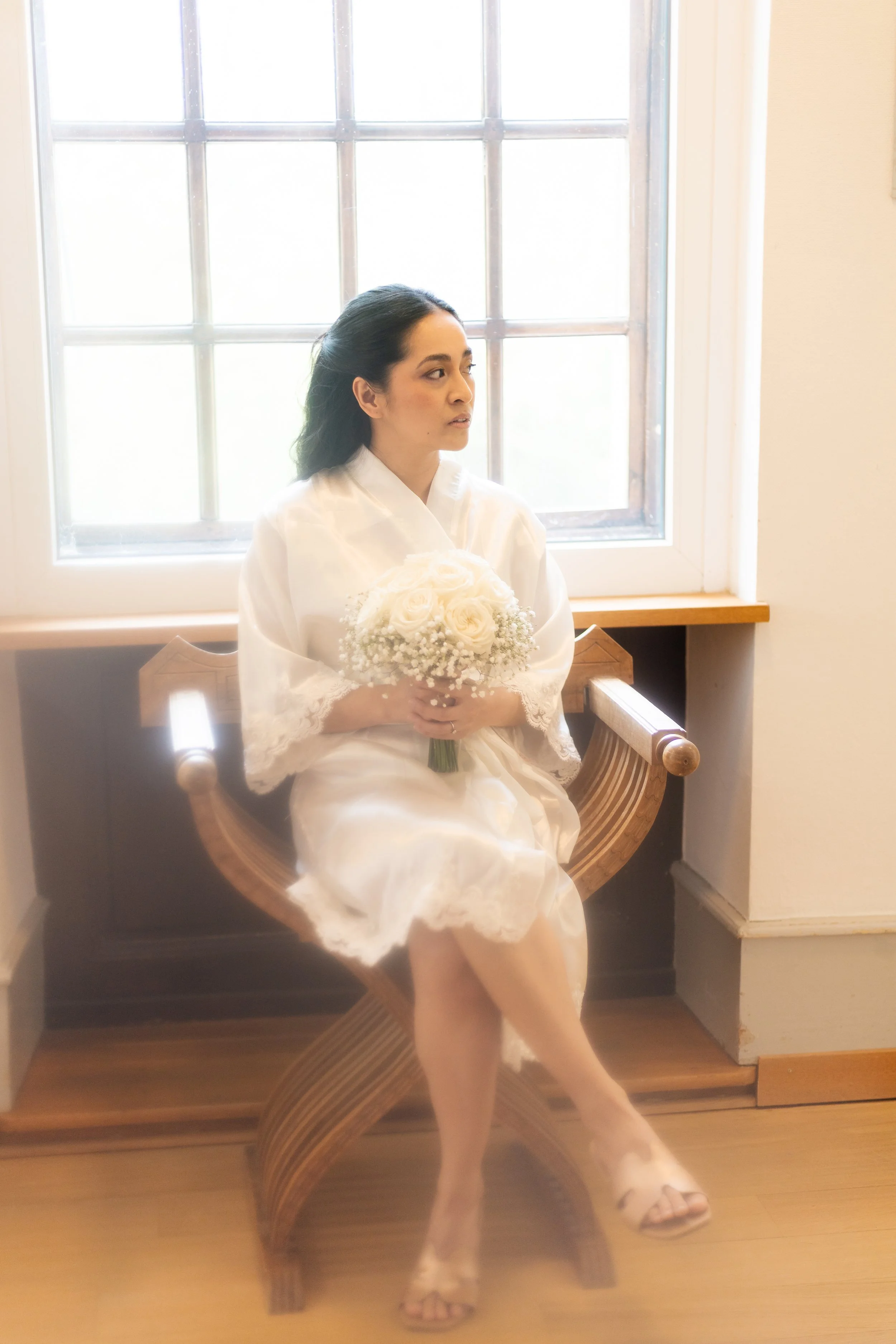 Une femme assise sur une chaise en bois, tenant un bouquet de fleurs blanches, dans une pièce éclairée par une grande fenêtre.