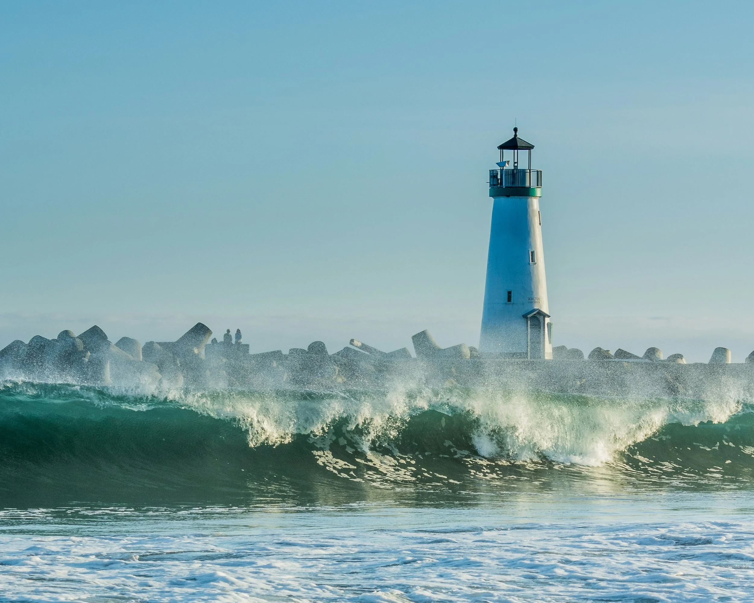 A tall white lighthouse with a black roof, situated near the shoreline with large rocks at its base, overlooking the ocean with waves and a clear sky. Descriptive to energy cleanse offered, as physical objects as well save and carry past energy.