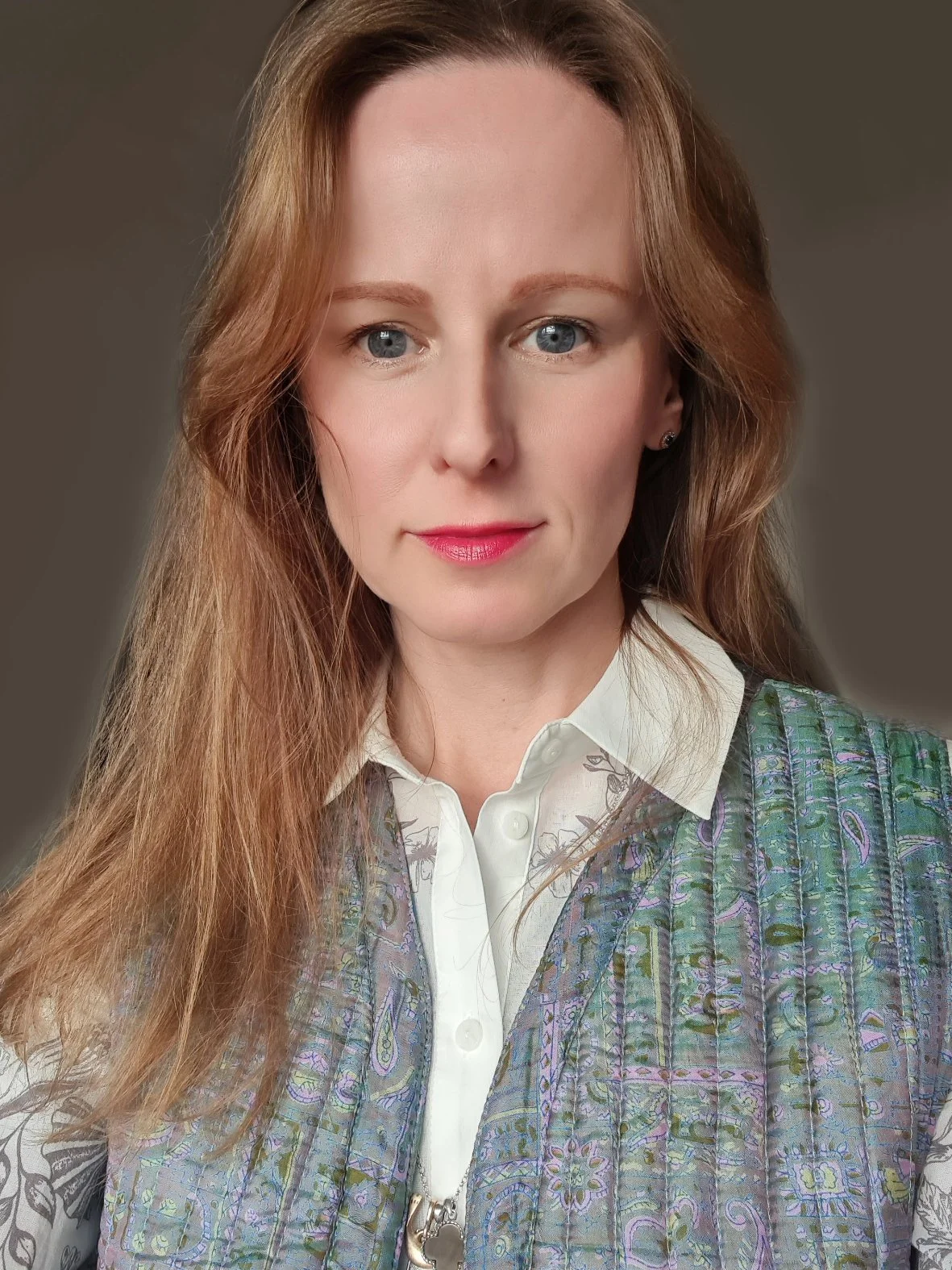 Portrait of a woman with long, light brown hair, blue eyes, wearing a white shirt and a patterned vest. Energy healing practitioner, hypnotherapist.