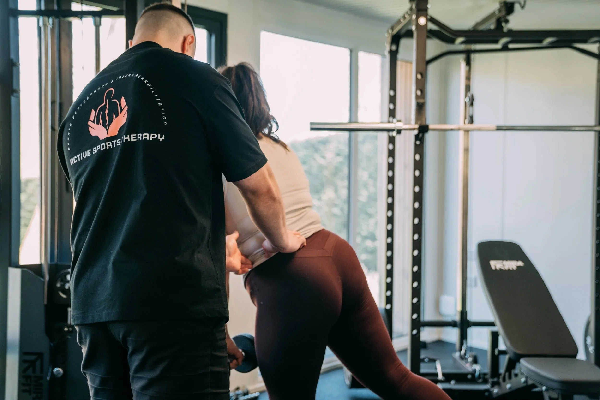 A personal trainer assists a woman in performing an exercise using gym equipment in a well-lit fitness center.