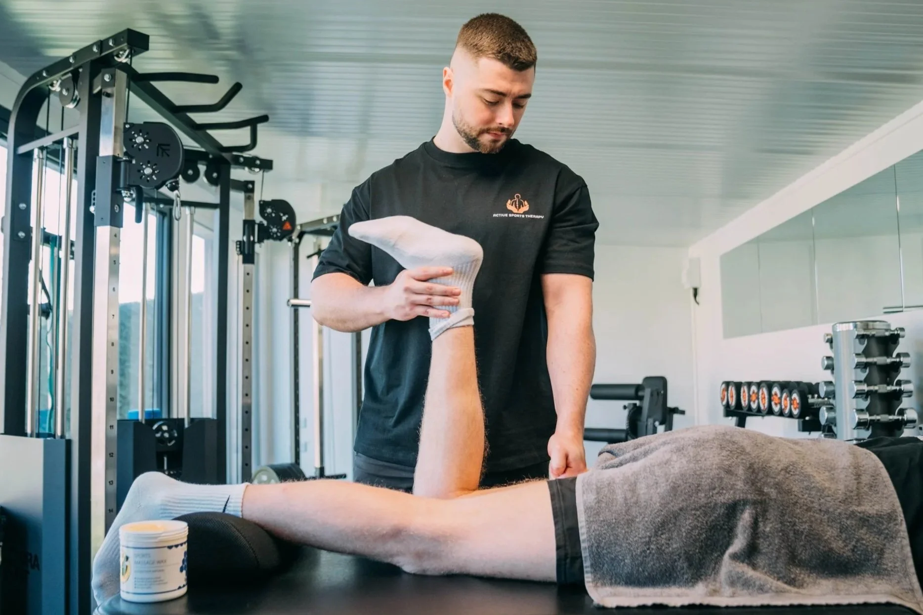 A physical therapist or sports therapist performs a leg stretch or rehab exercise on a client lying on a treatment table in a gym or therapy center, with exercise equipment visible in the background.
