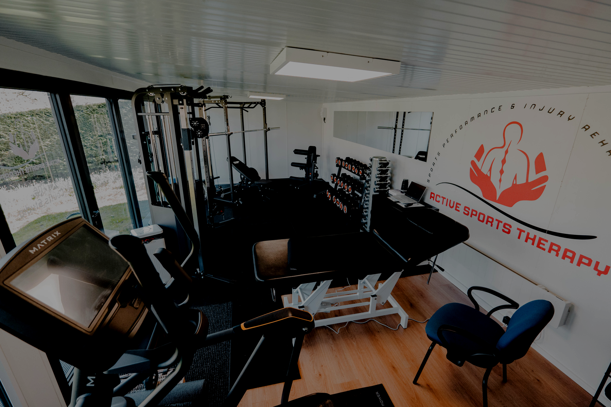 Small gym room with workout equipment including dumbbells, weightlifting stations, a treadmill, on a wood floor and a wall with the logo and name of a sports therapy business, large windows with outdoor greenery.