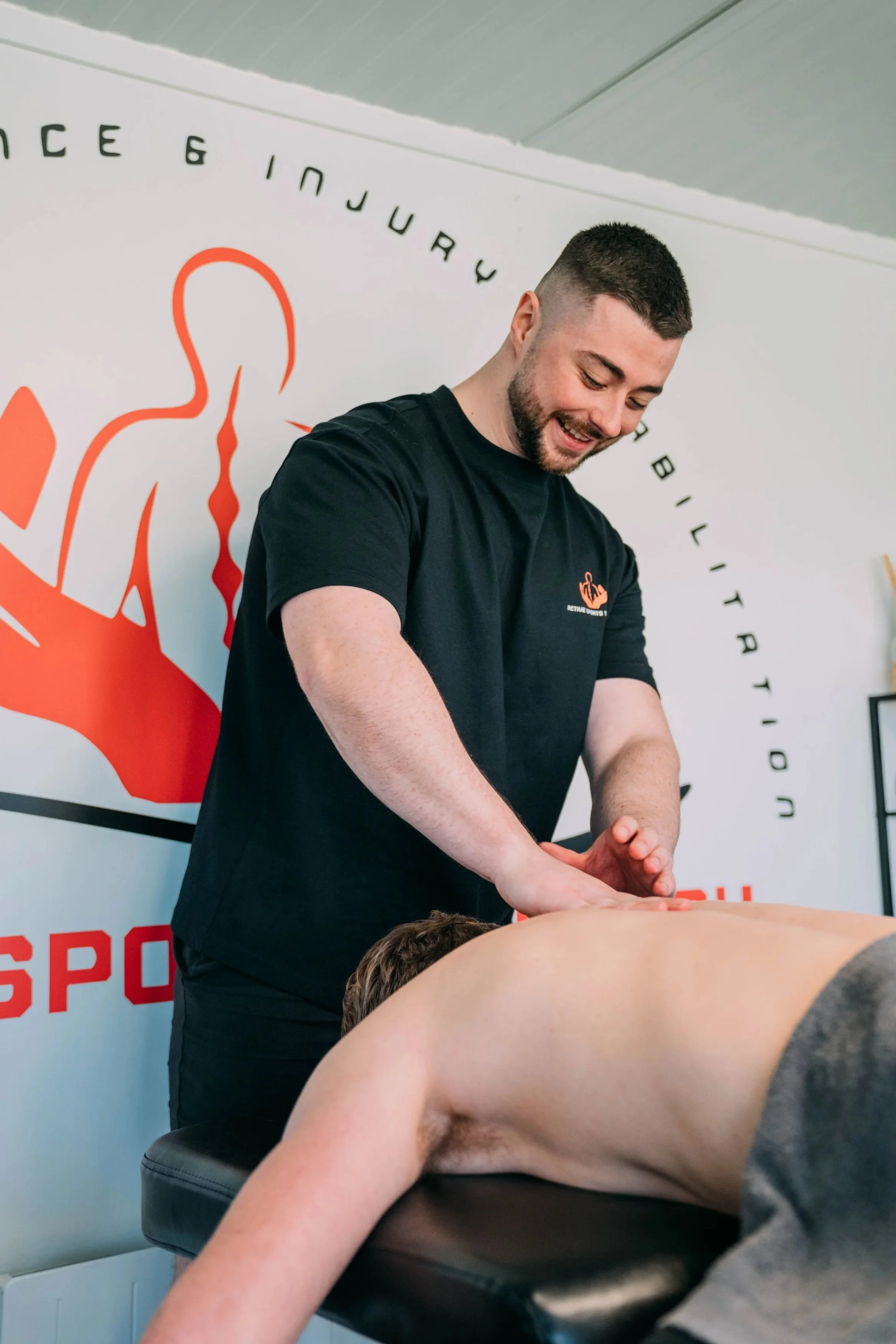 A man receiving a massage in a clinic or spa. The man is lying face down on a massage table while a massage therapist applies pressure to his back. The background shows a wall with a logo featuring an abstract human figure and some text.