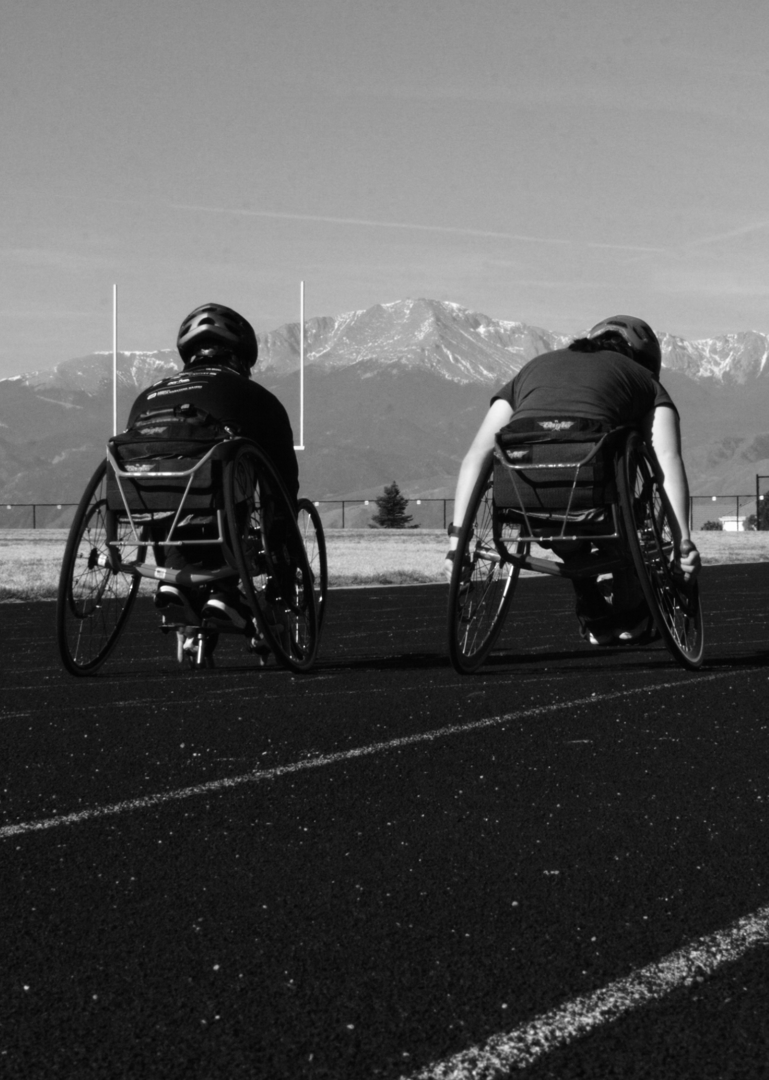 Two individuals in wheelchairs on an outdoor track, facing away and looking at mountains in the distance, with a football goal post and clear sky.