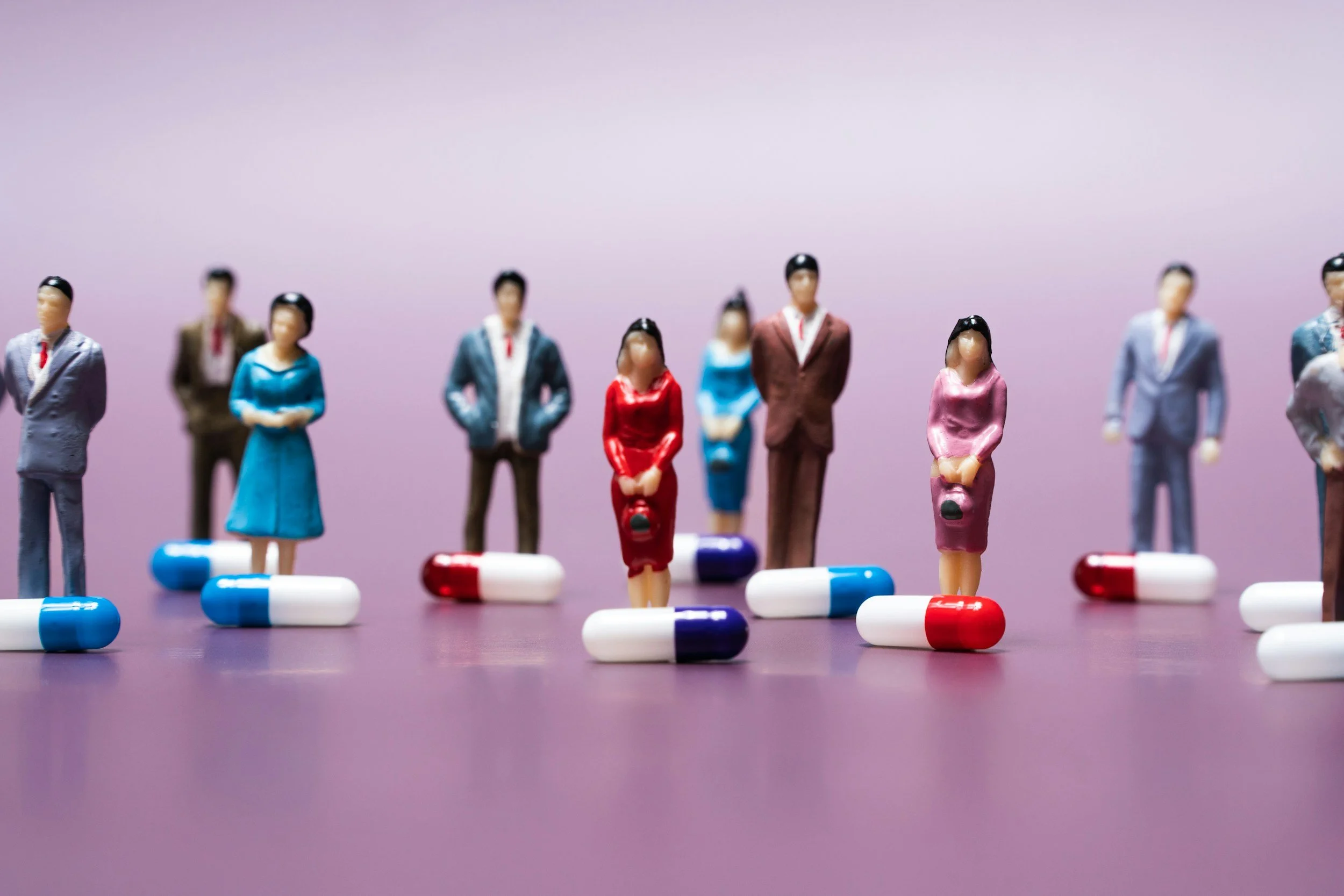 Miniature figurines of people in business attire standing on top of pills, set against a purple background.