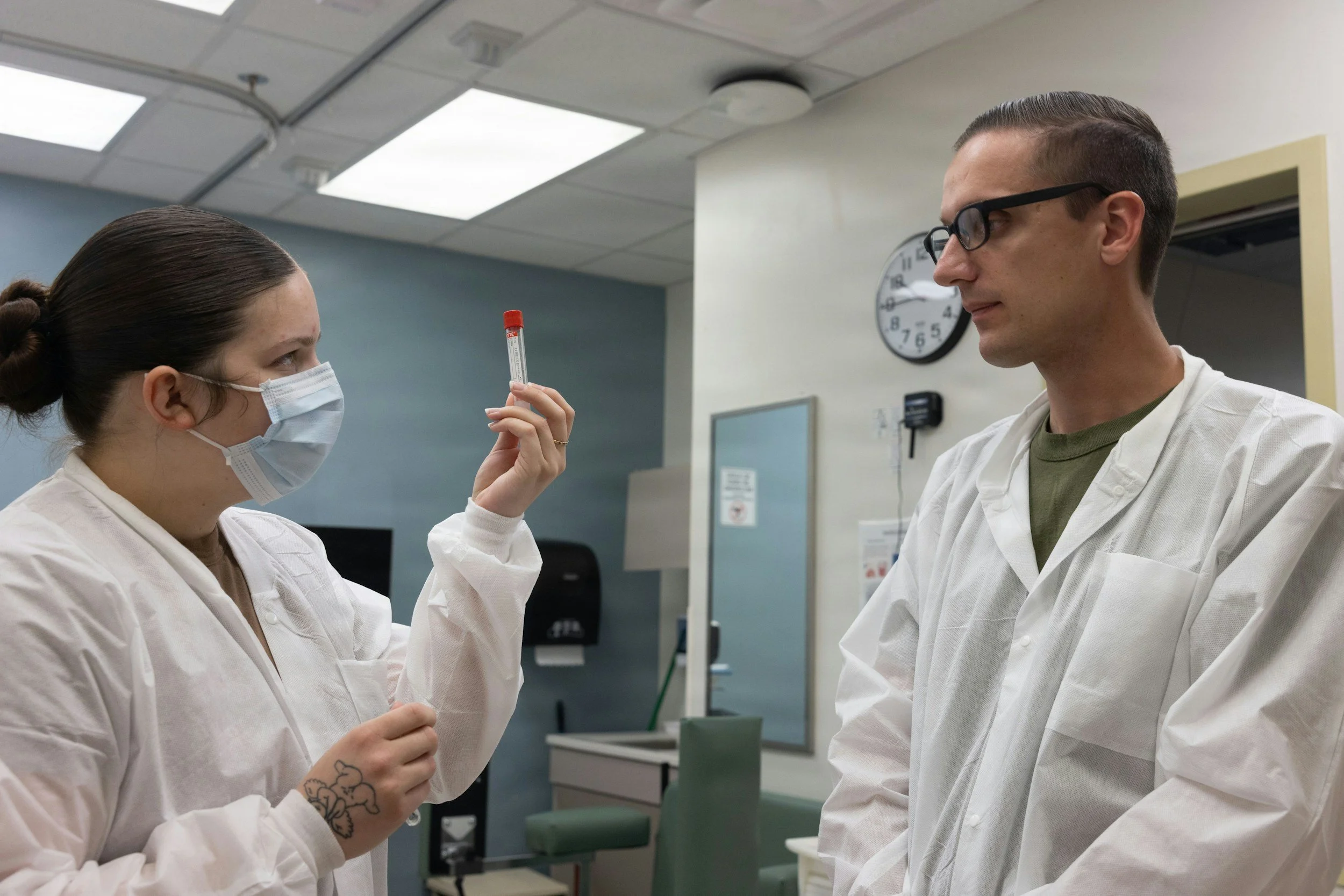 A female healthcare worker wearing a face mask and lab coat holding a test tube with a red cap, speaking with a male healthcare worker wearing glasses and a lab coat in a medical setting.