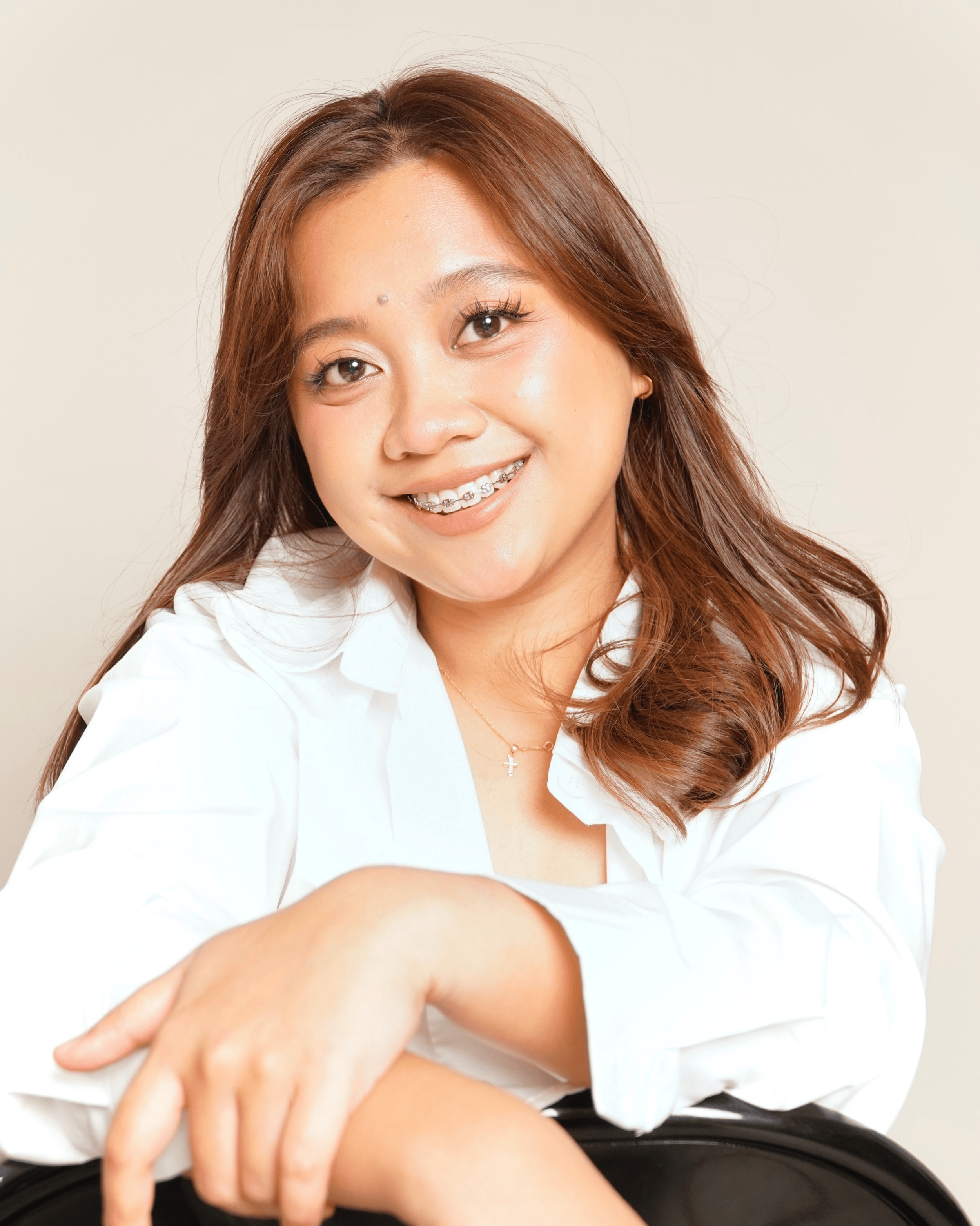 A young woman with long, wavy brown hair smiling, wearing braces and a white blouse with rolled-up sleeves, sitting with her arm resting on a black chair.