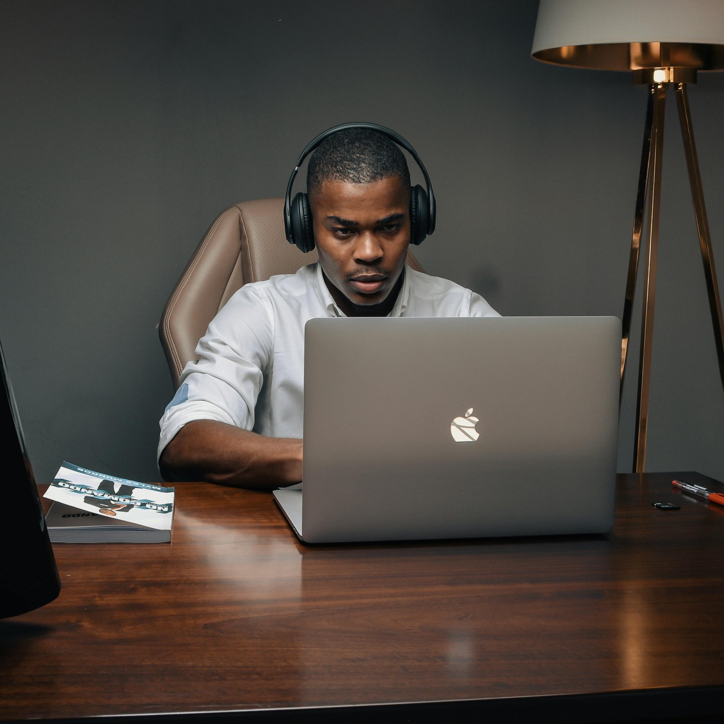 Young man working at a desk with a laptop, headphones, and a magazine in a modern office setting.