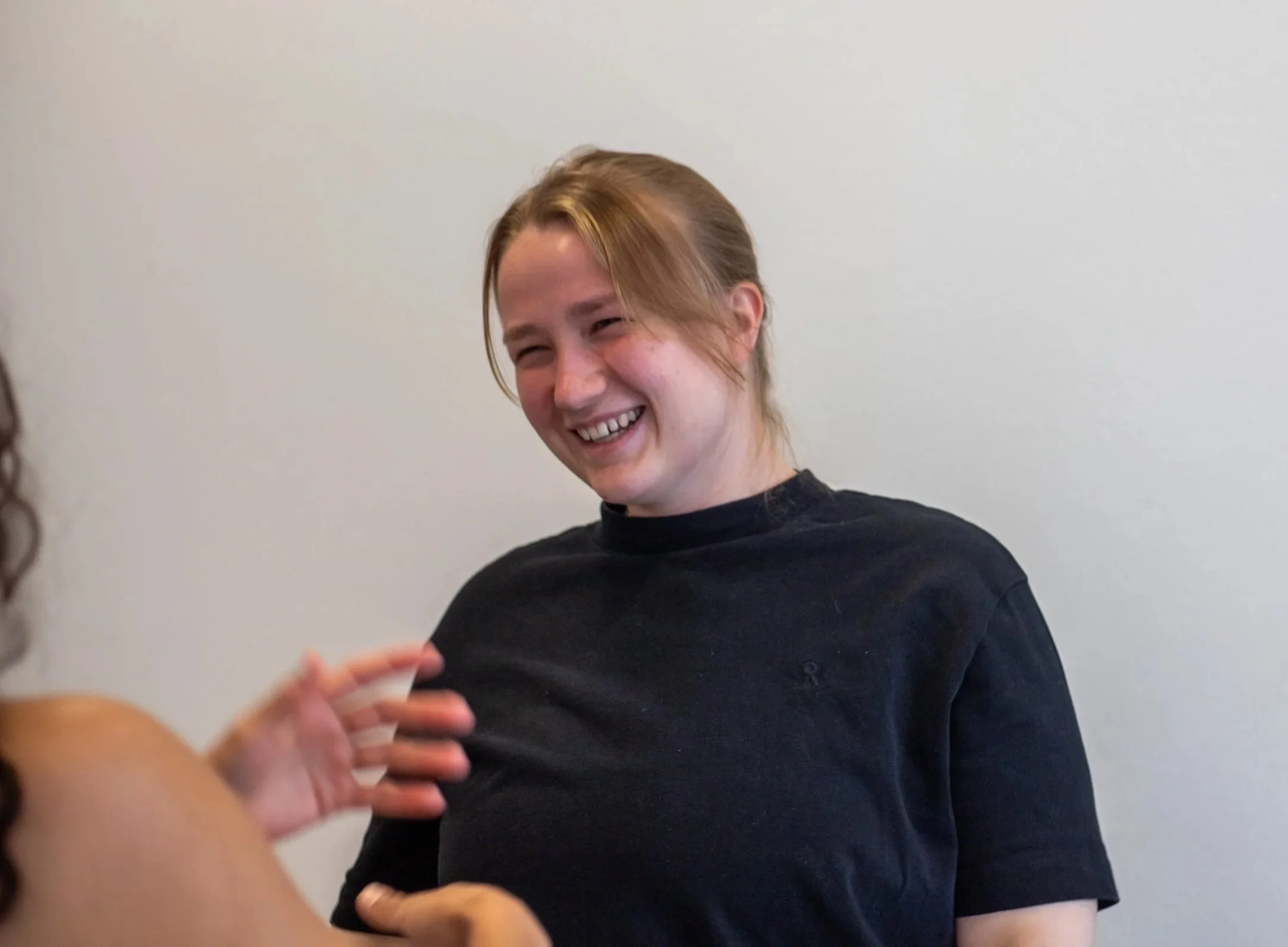 A young woman with light brown hair in a ponytail, wearing a black shirt, smiling and looking at another person, against a plain white wall.
