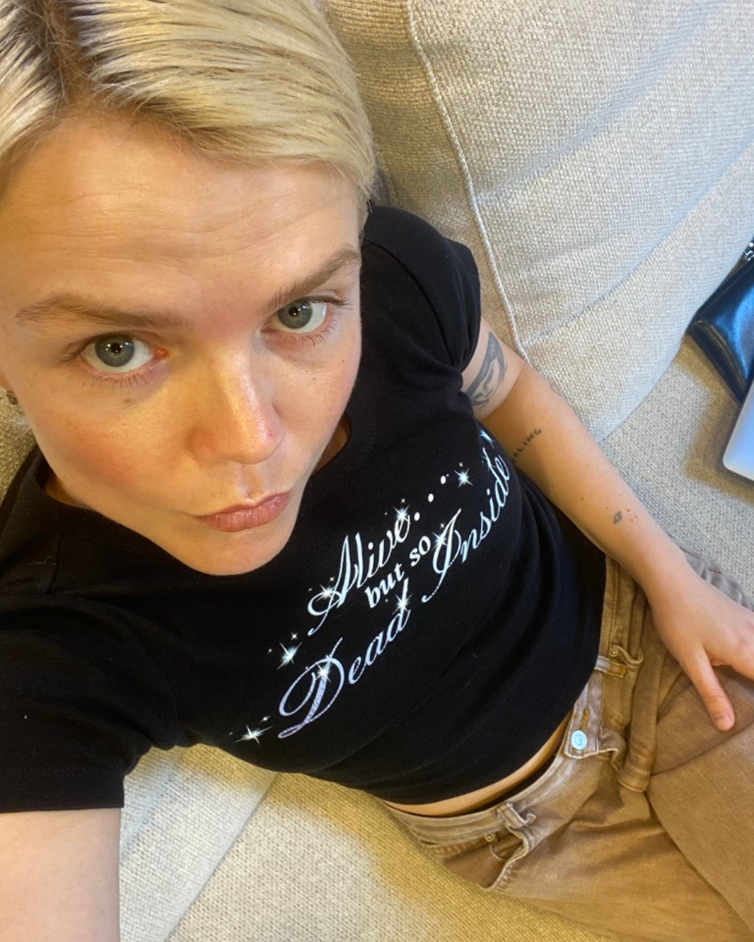 En person med ljus blond frisyr, tittar upp mot kameran, liggande på en beige soffkudde, bär en svart T-shirt med vit text och beigea byxor.