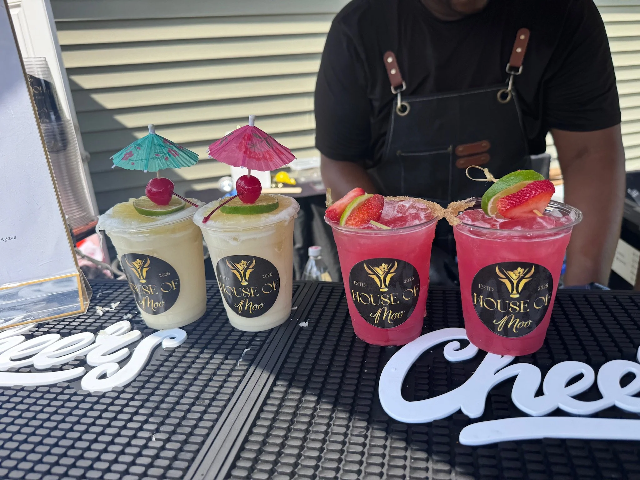 Four colorful beverages in cups topped with lime slices, cherries, and miniature umbrellas, placed on a black table with decorative signage.