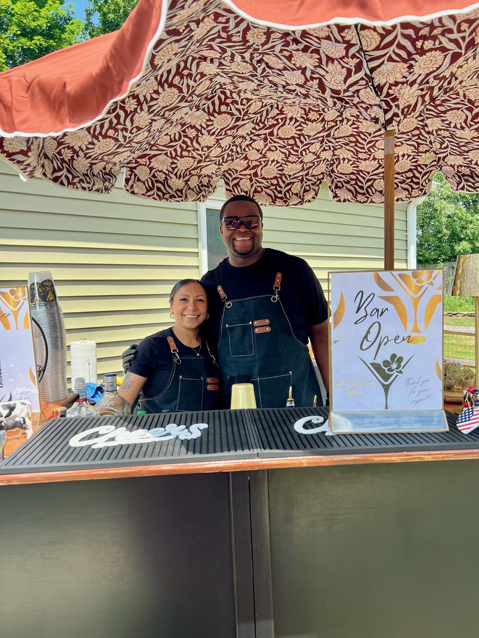 young entrepreneurs minority husband and wife standing in front of black bar with brown top bar open sign bar mats with the word cheers with floral umbrella over them