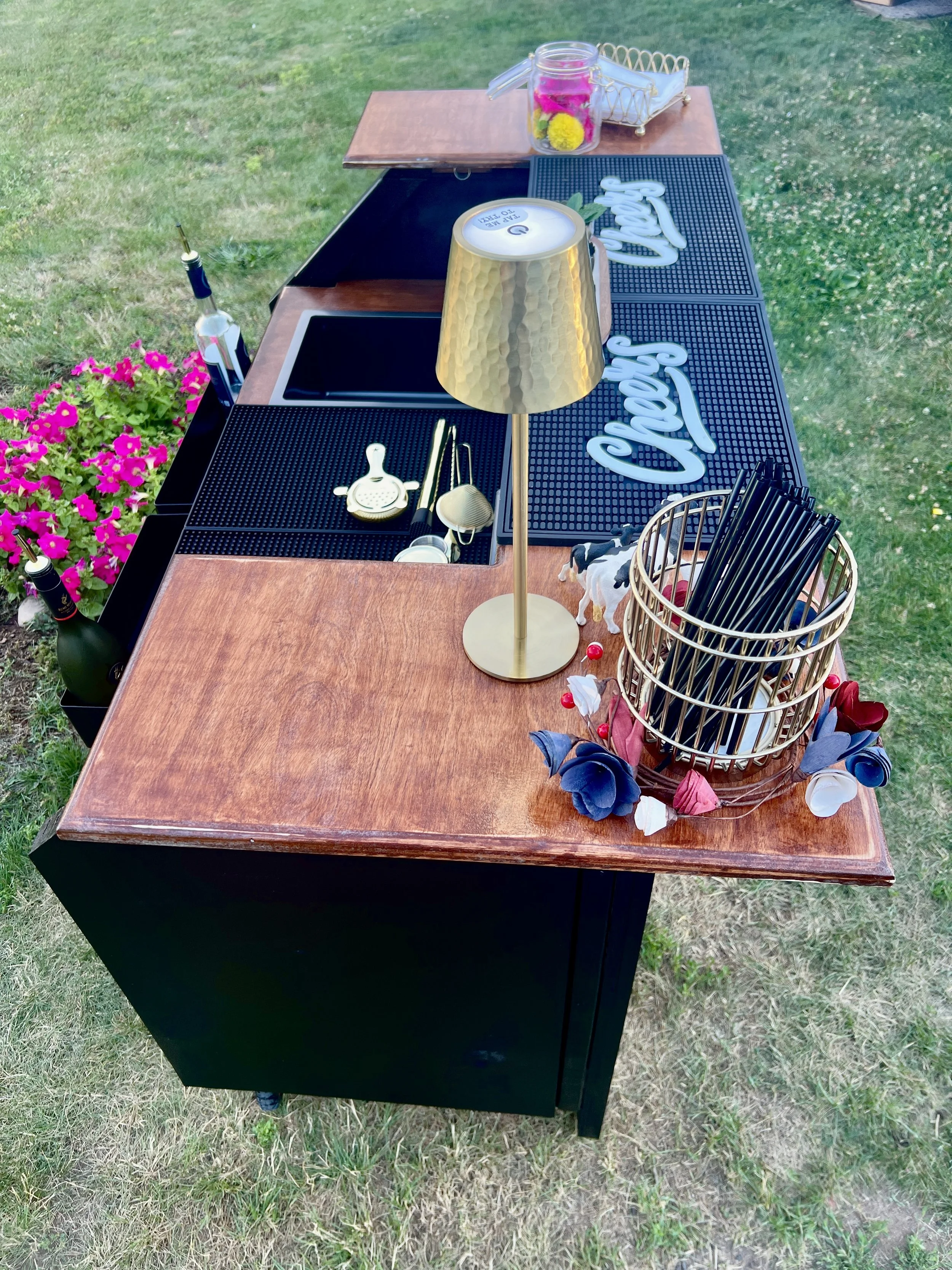 A fully equipped portable mobile bar station set up outdoors on grass. The wooden bar top is organized with a sleek gold lamp, black rubber spill mats featuring white "Cheers" text, a gold wire basket of straws, a cocktail strainer, a built-in ice or