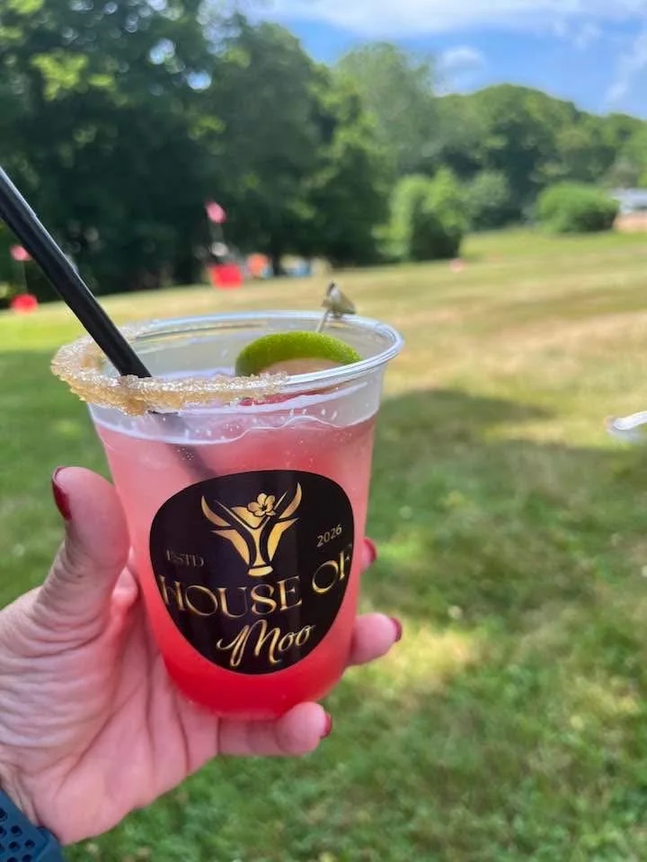 A hand holding a pink cocktail with a lime wedge and a sugar-rimmed edge, in a plastic cup labeled "House of Moo." In the background, there are trees and an outdoor grassy area under a partly cloudy sky.