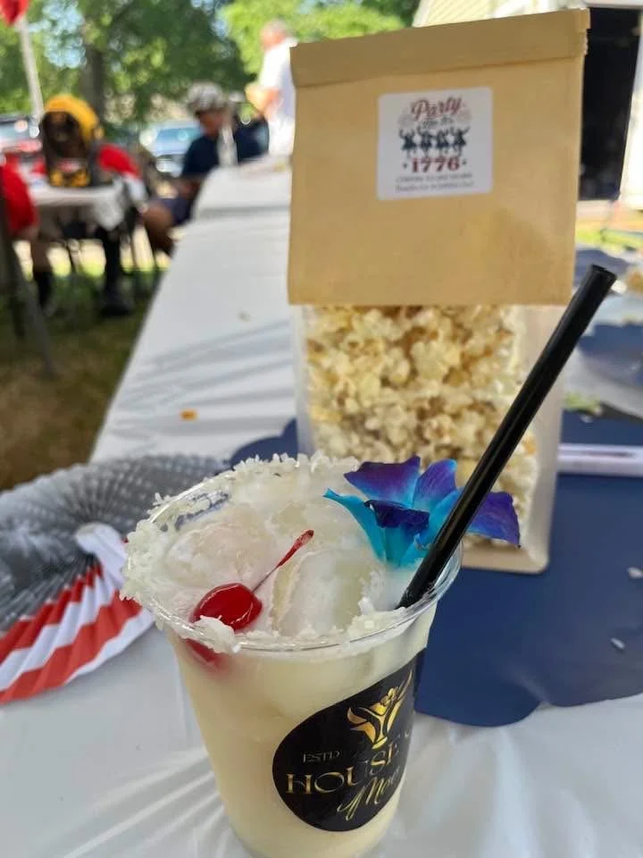 A tropical cocktail with a cherry and blue flower garnish on ice, in a clear cup with a black straw, on a table at an outdoor event. In the background, there is a bag of popcorn and people sitting at tables.