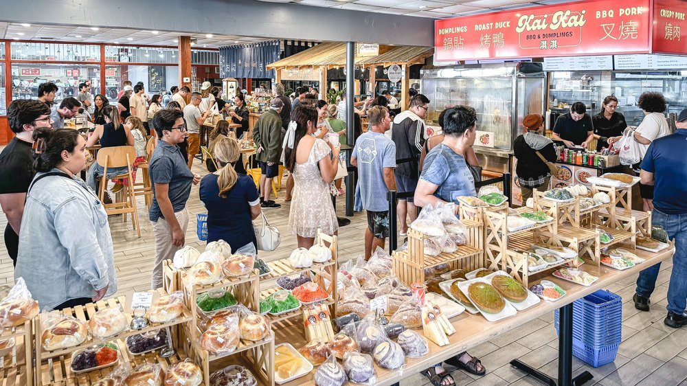 Crowded indoor marketplace with many people shopping and browsing food stalls, including a bakery section with various baked goods, and a food stand selling BBQ and pork dishes.