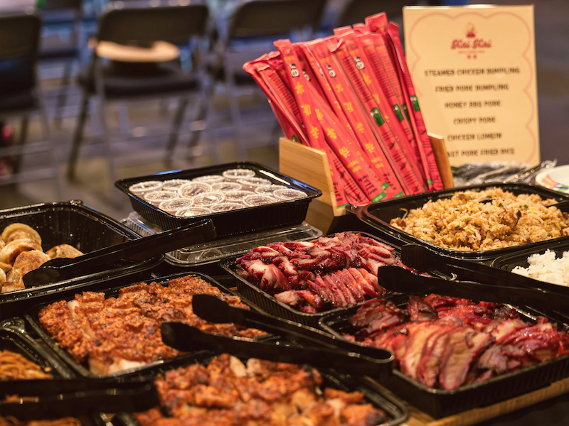 Kai Kai BBQ catering with various dishes including sliced roasted meat, rice, and side dishes, along with chopsticks and a menu in the background.