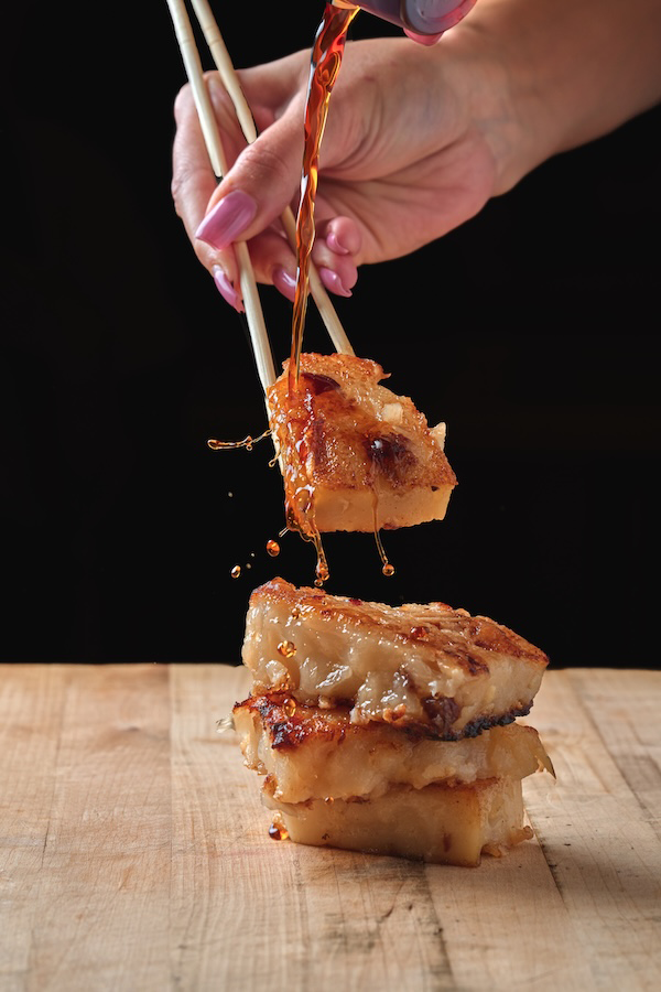 Hand holding chopsticks picking up a piece of Kai Kai BBQ fried tofu with soy sauce being poured over it.