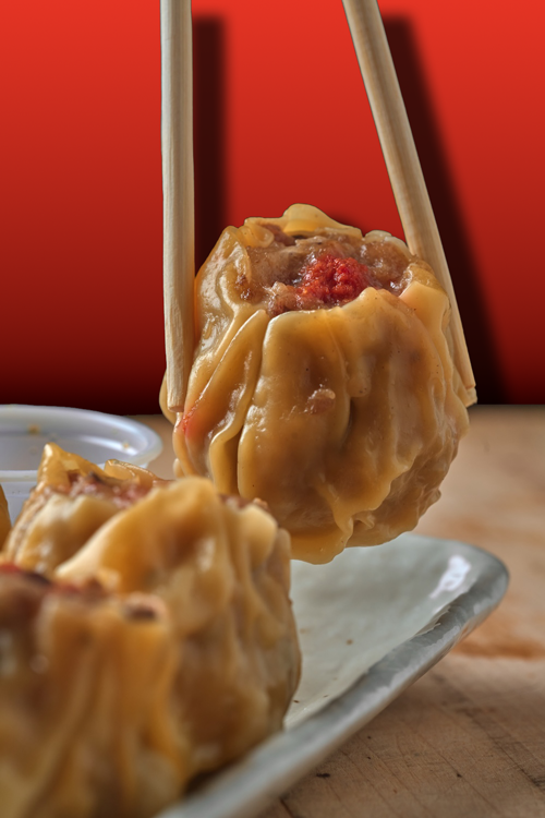 Close-up of a Kai Kai BBQ Siu Mai dumpling held with chopsticks over a white plate, with another dumpling on the plate, against a red background.