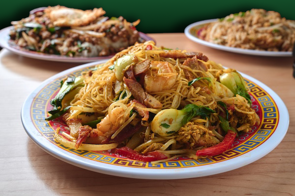 A plate of Kai Kai BBQ stir-fried noodles with vegetables, shrimp, and sliced meat, with two other dishes in the background.