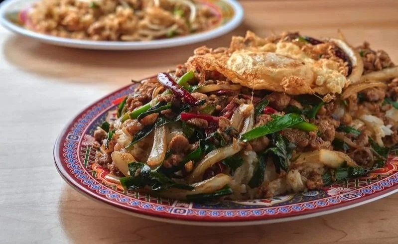 Plate of Kai Kai BBQ stir-fried ground meat with vegetables and a fried egg, with a second dish of noodles in the background.