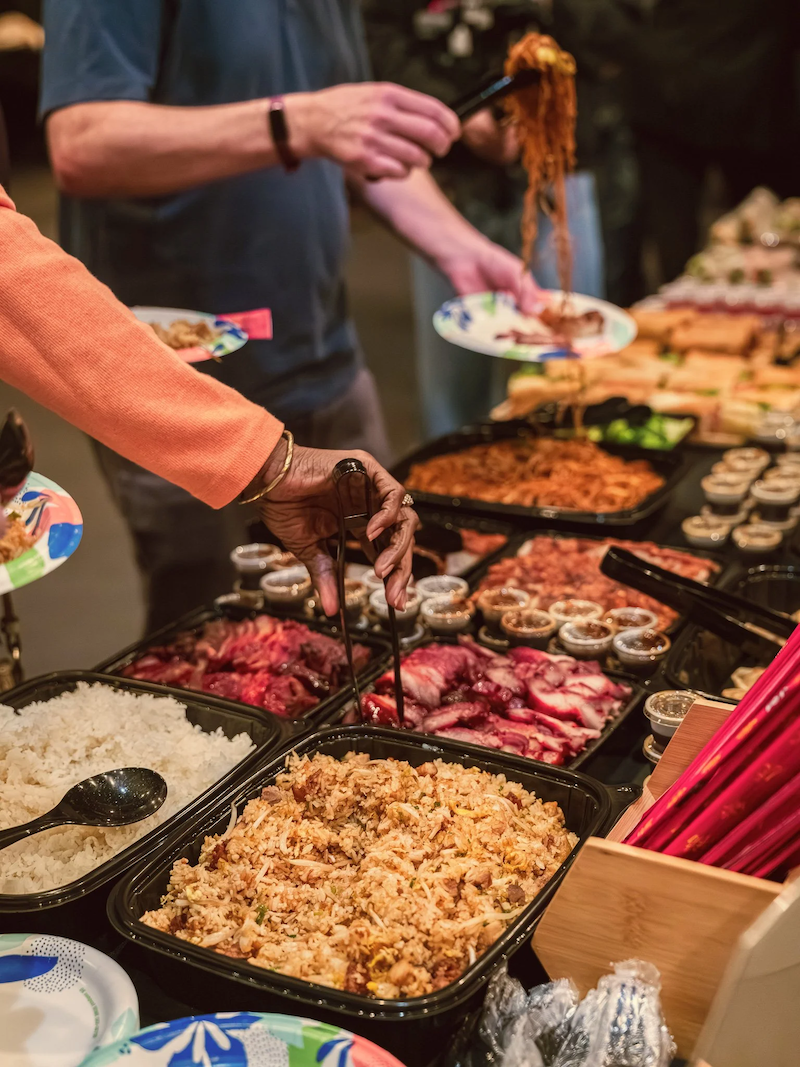 Kai Kai BBQ catering with various dishes including sliced roasted meat, rice, and side dishes, along with chopsticks and a menu in the background.