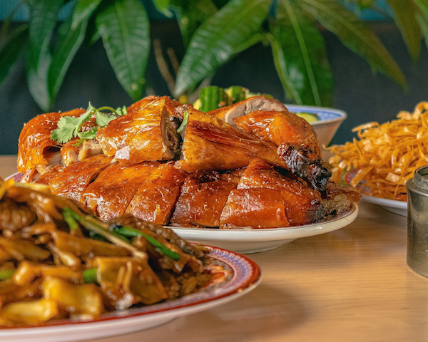 Platter of roasted and sliced chicken with a garnish of cilantro, accompanied by stir-fried vegetables and noodles on separate plates, with greenery in the background.