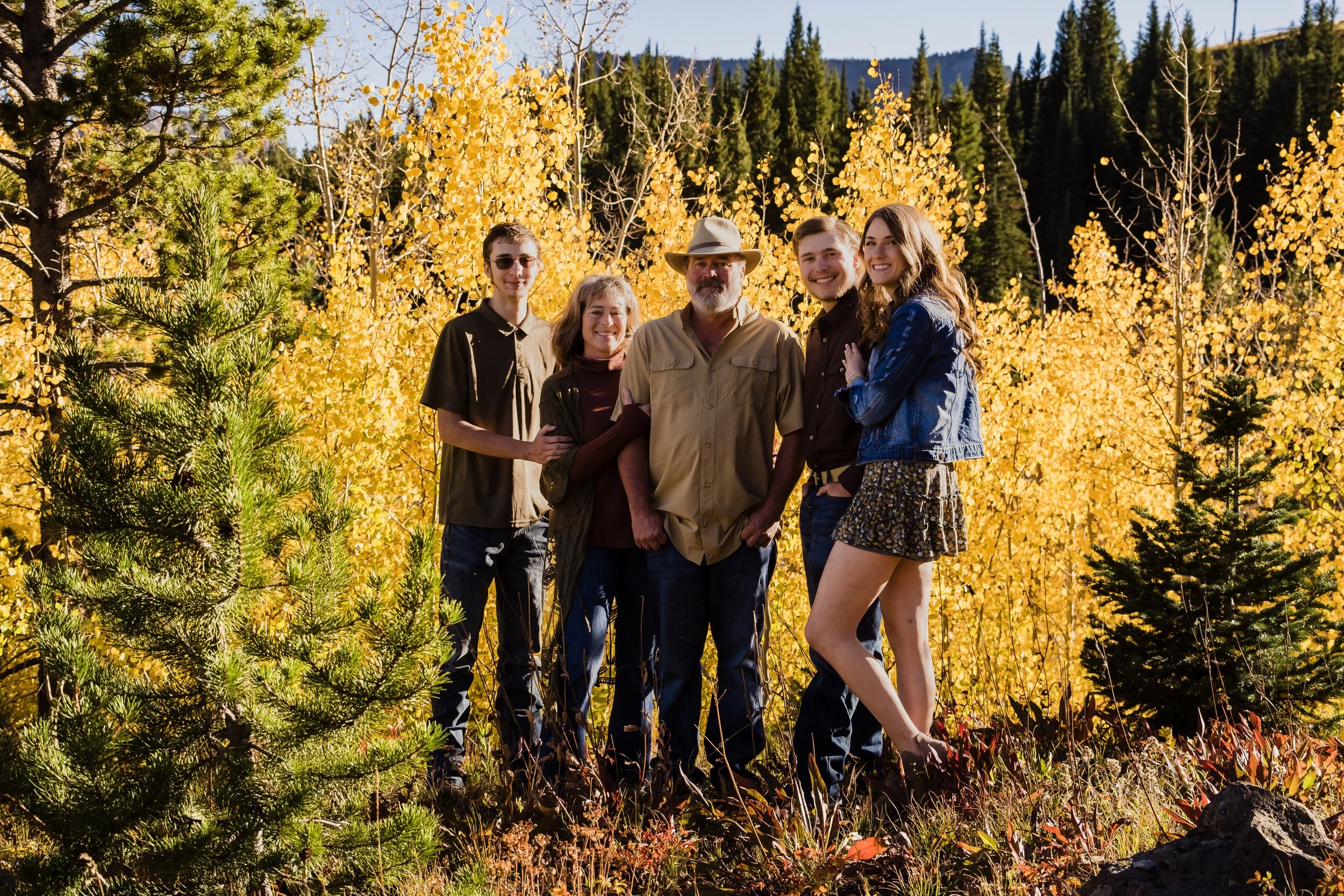 Five people smiling and standing together outdoors in a forest with autumn-colored trees in the background.