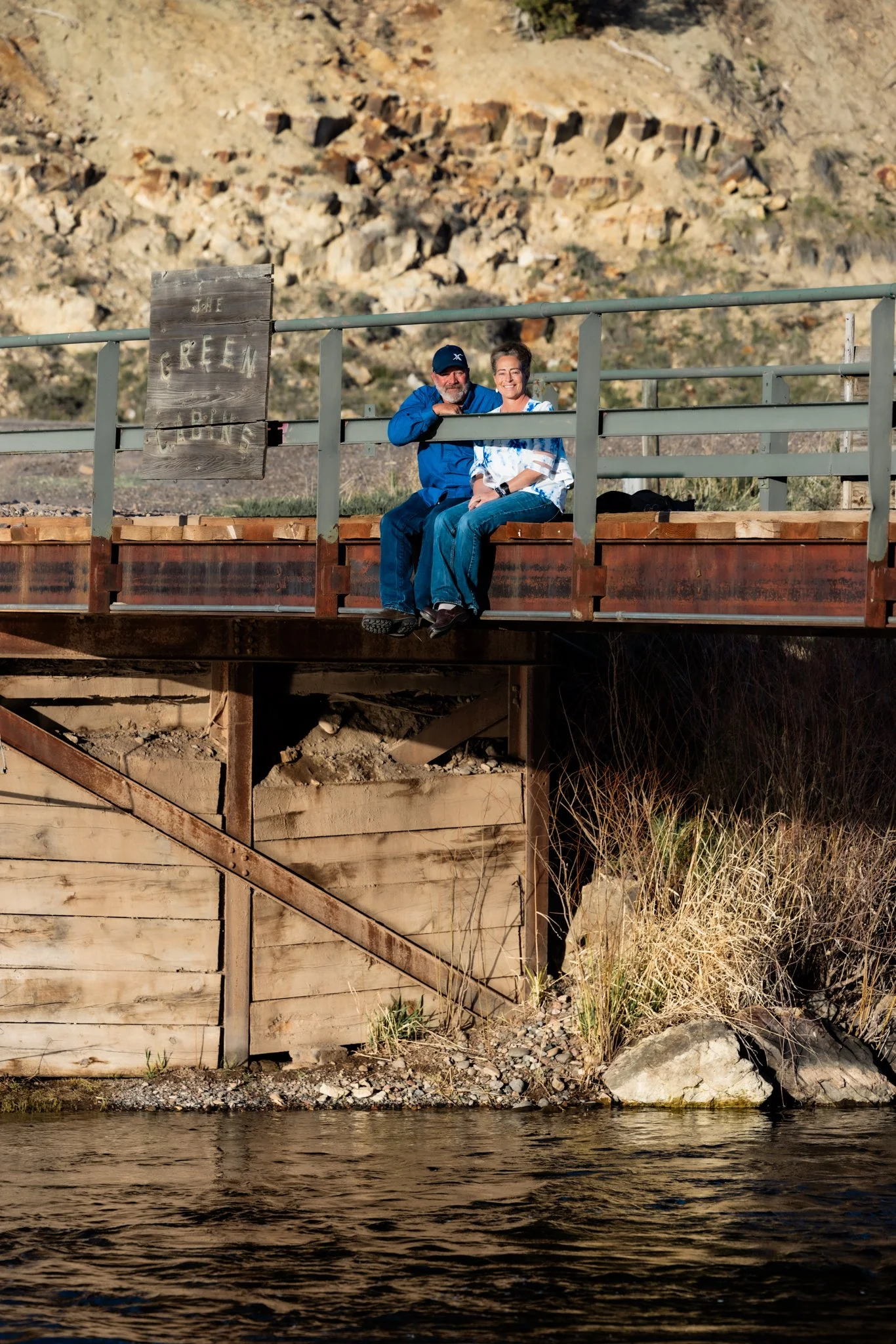 Two people sitting on a wooden and metal bridge over a river, smiling and enjoying the outdoors.