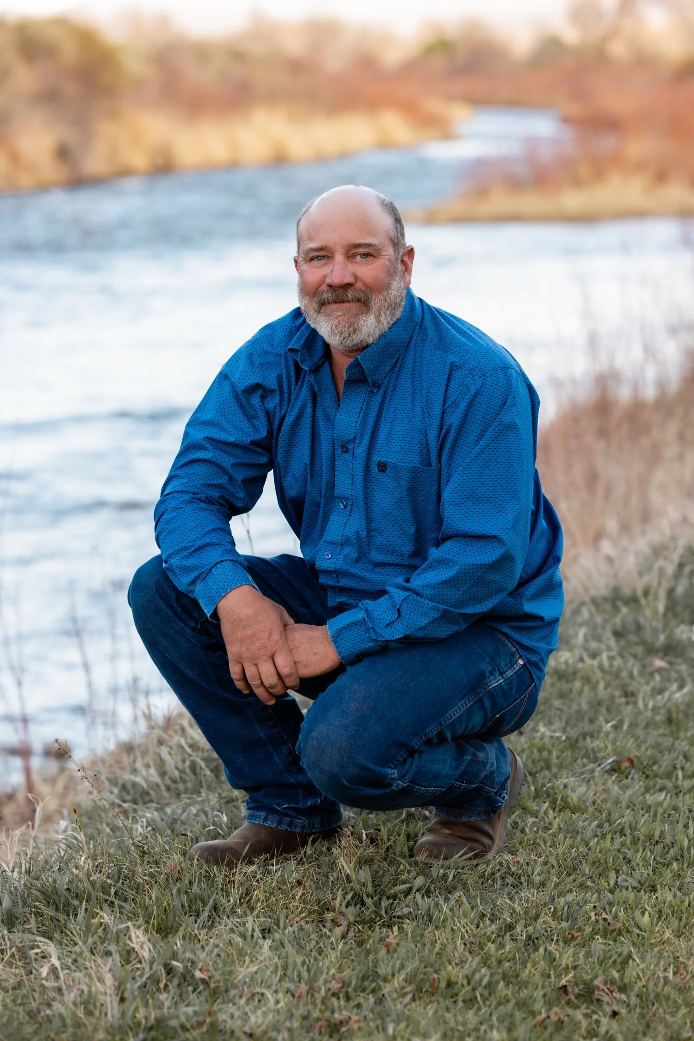 A man with a beard kneeling on grass near a river during daytime, wearing a blue button-up shirt and jeans.