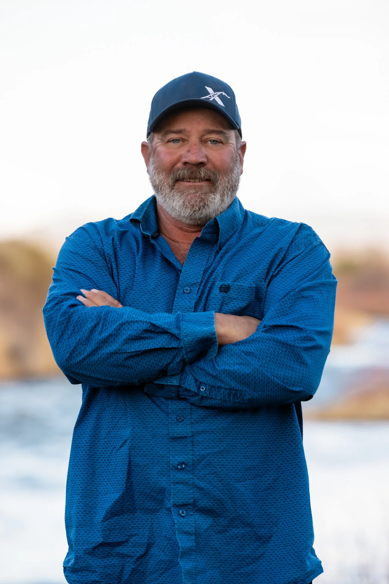 A man with a gray beard and mustache, wearing a navy blue baseball cap and a blue button-up shirt, stands outdoors with arms crossed, blurred natural background.