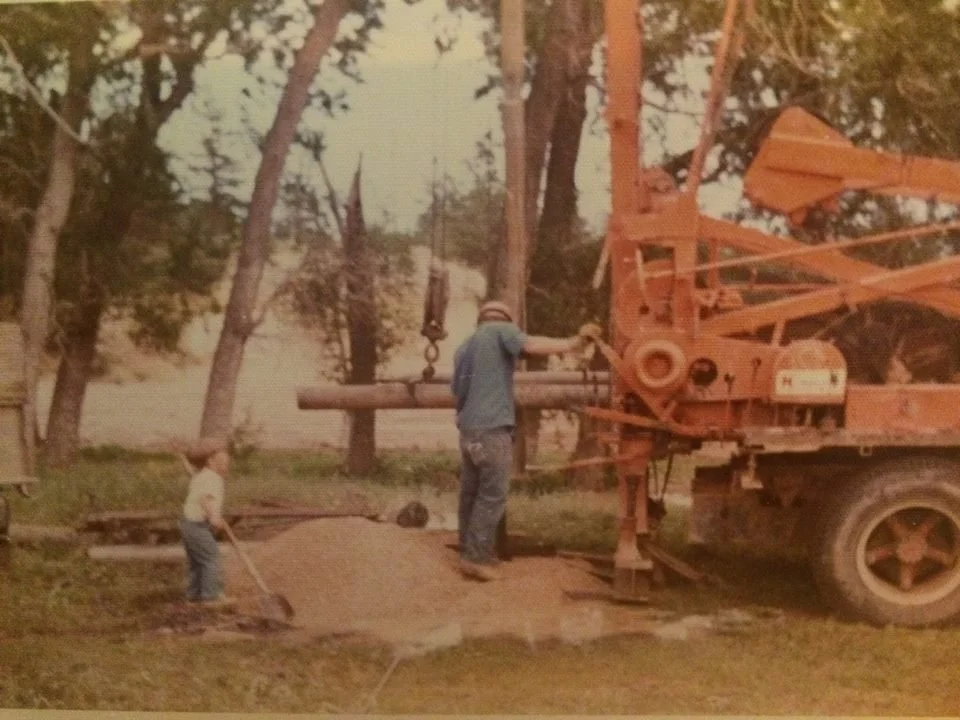 2 generations of Day's operating a large orange construction drill on a grassy area with trees in the background.