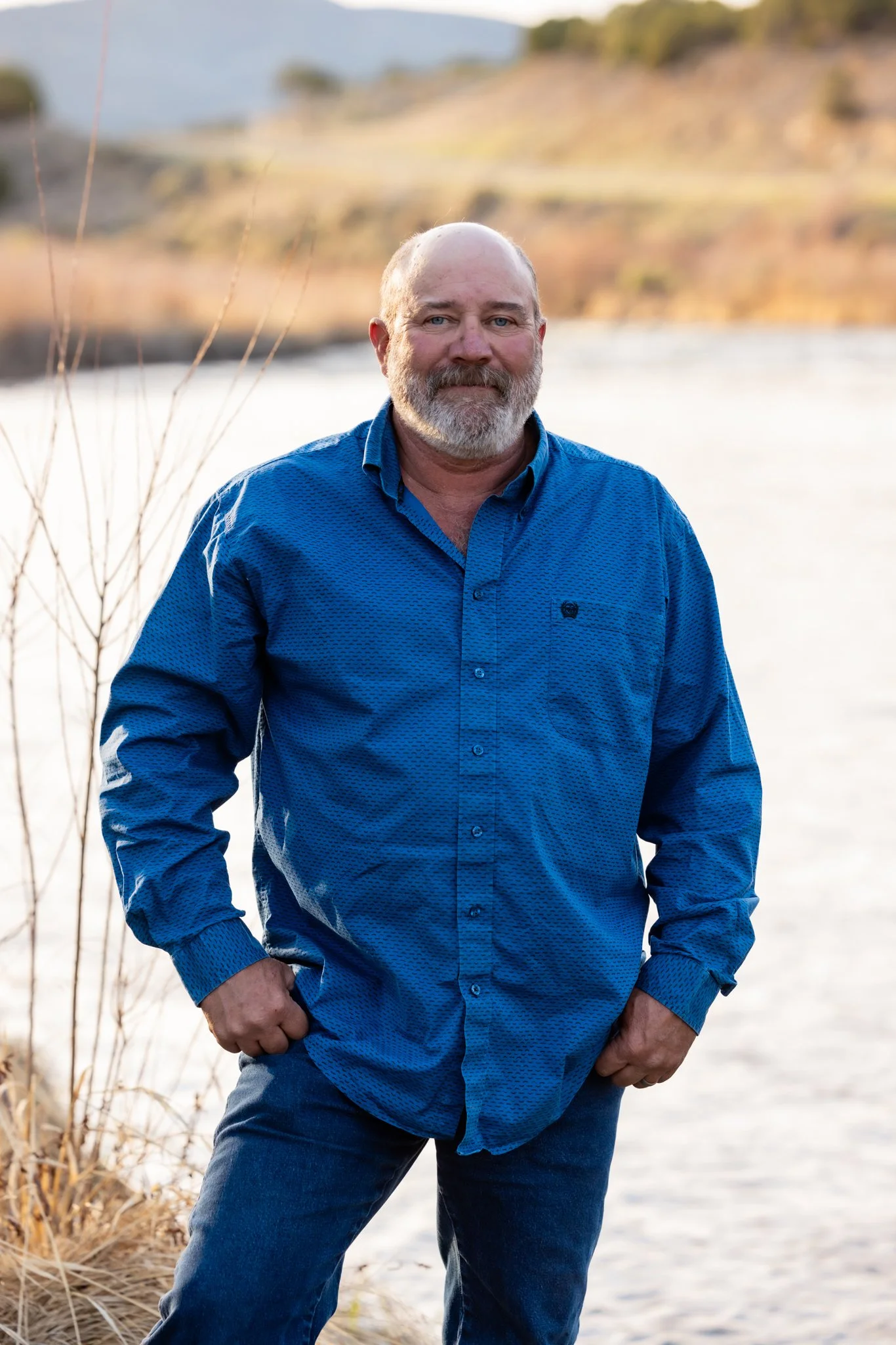 A man with a beard wearing a blue shirt and dark jeans standing outdoors near a body of water with a hillside in the background.