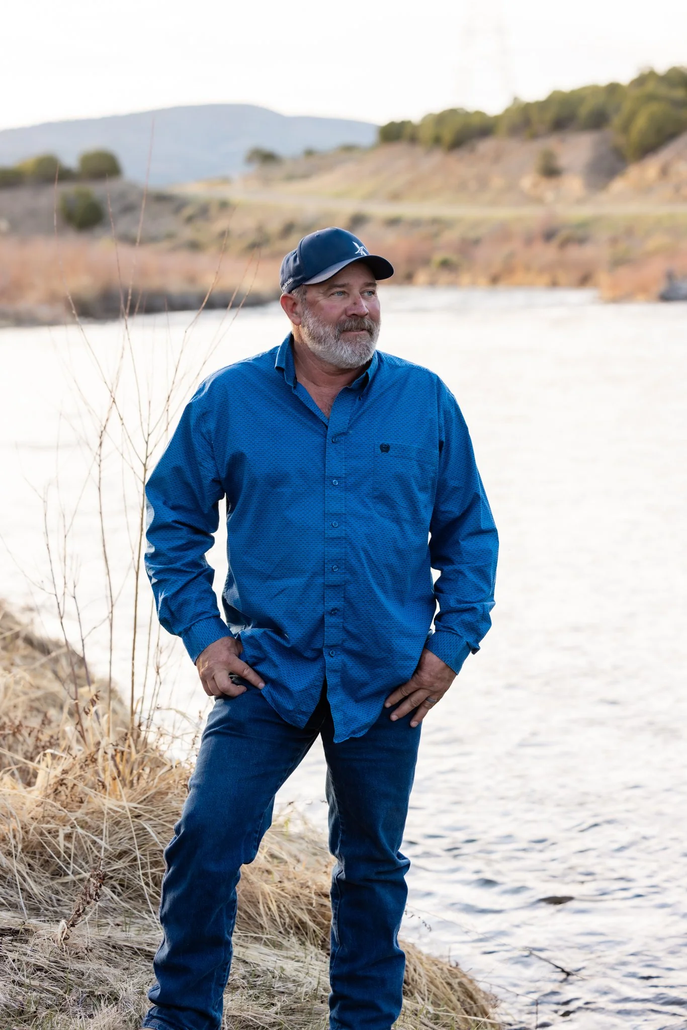 A man with a beard wearing a blue button-up shirt, jeans, and a baseball cap standing by a river with hands in pockets, overlooking water and distant hills.