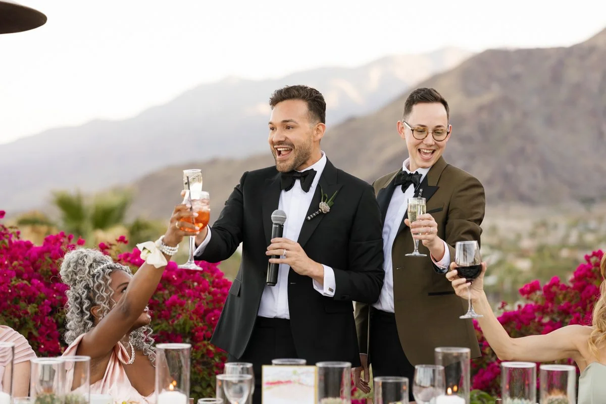 Two men in tuxedos celebrating with drinks at an outdoor wedding reception, with pink flowers and mountains in the background.