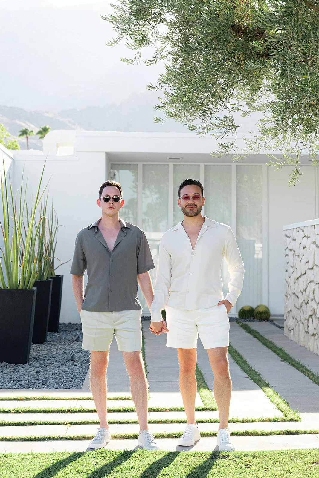 Two men standing outdoors in front of a modern white building, holding hands. Both wear sunglasses and casual summer outfits with shorts and open-collared shirts, one in grey and the other in white.