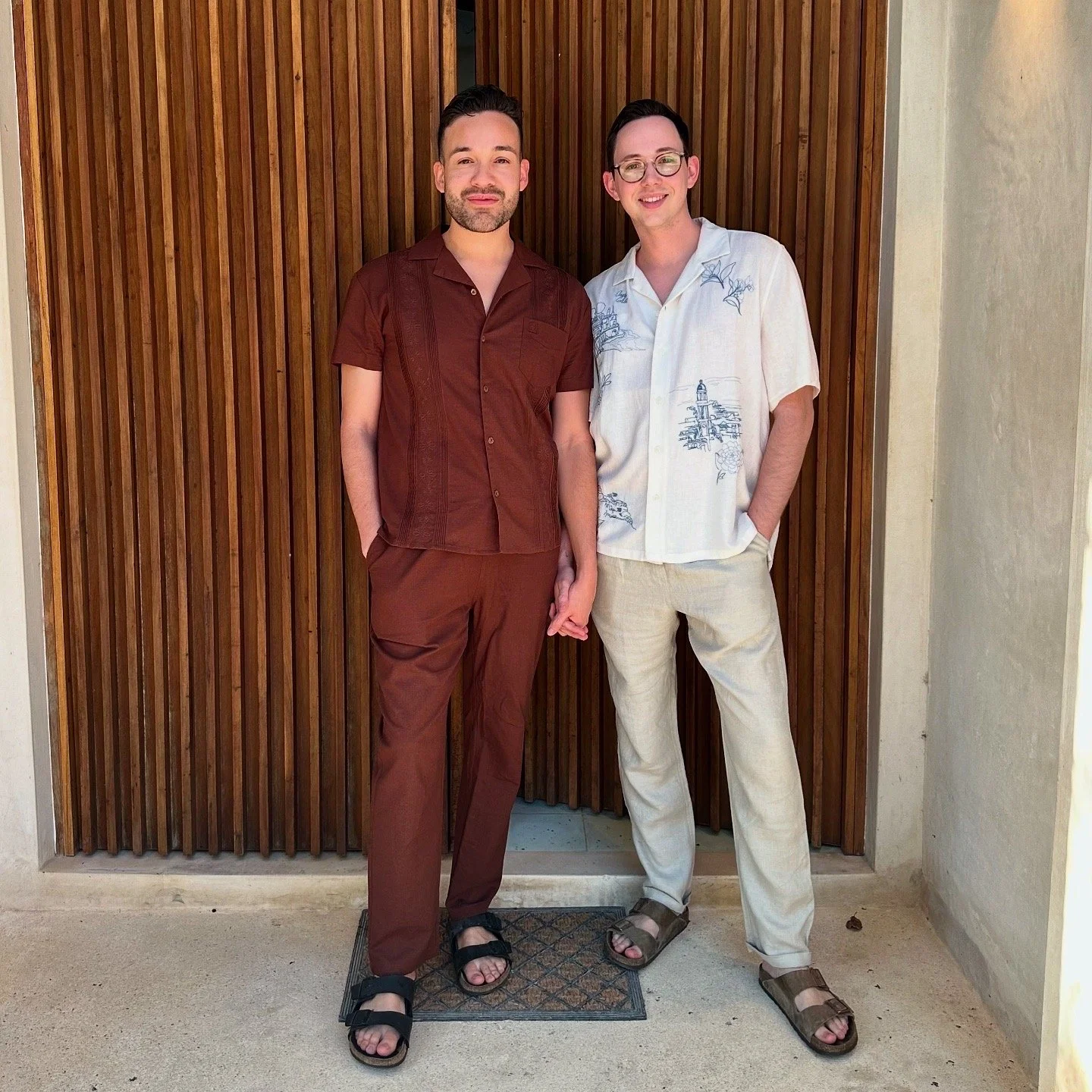Two men standing side by side, holding hands, in front of a wooden wall. One man is wearing a brown short-sleeved shirt and matching pants, and the other is wearing a white, short-sleeved, button-up shirt with a blue pattern and light-colored pants. They are both smiling.