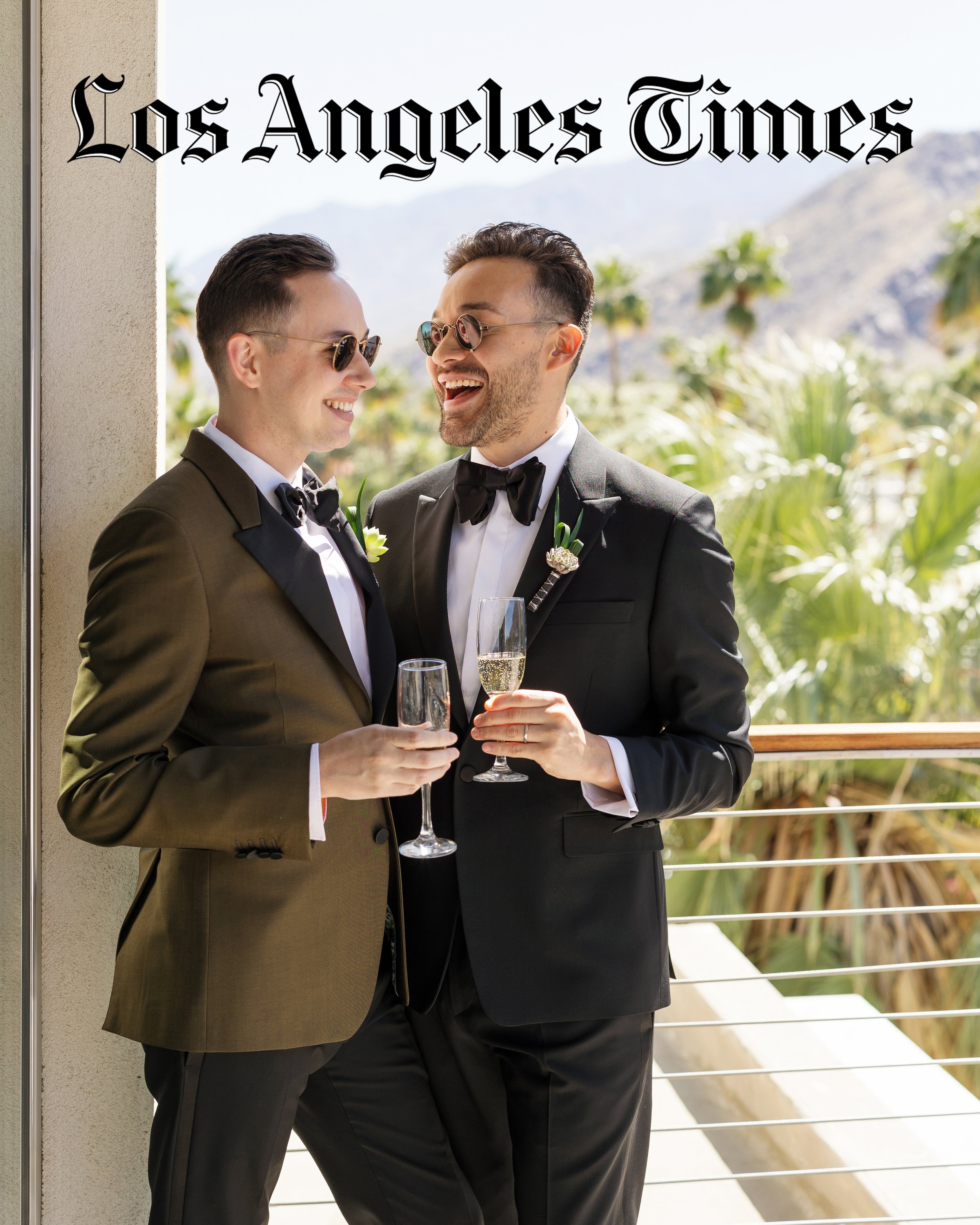 Two men in tuxedos holding champagne glasses, smiling and laughing on a balcony with palm trees and mountains in the background.