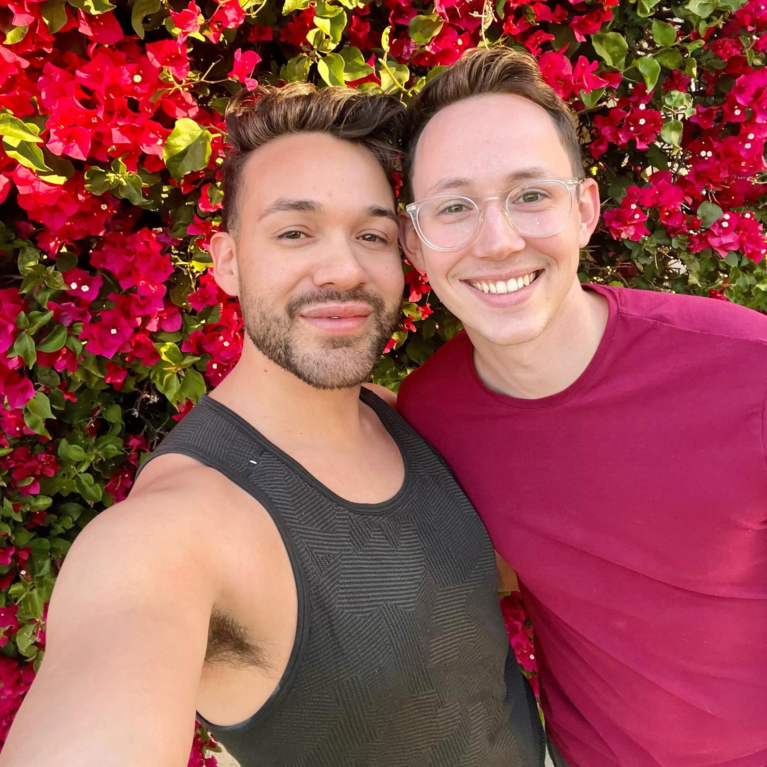 Two men smiling and taking a selfie in front of a vibrant pink and red flowering bush.