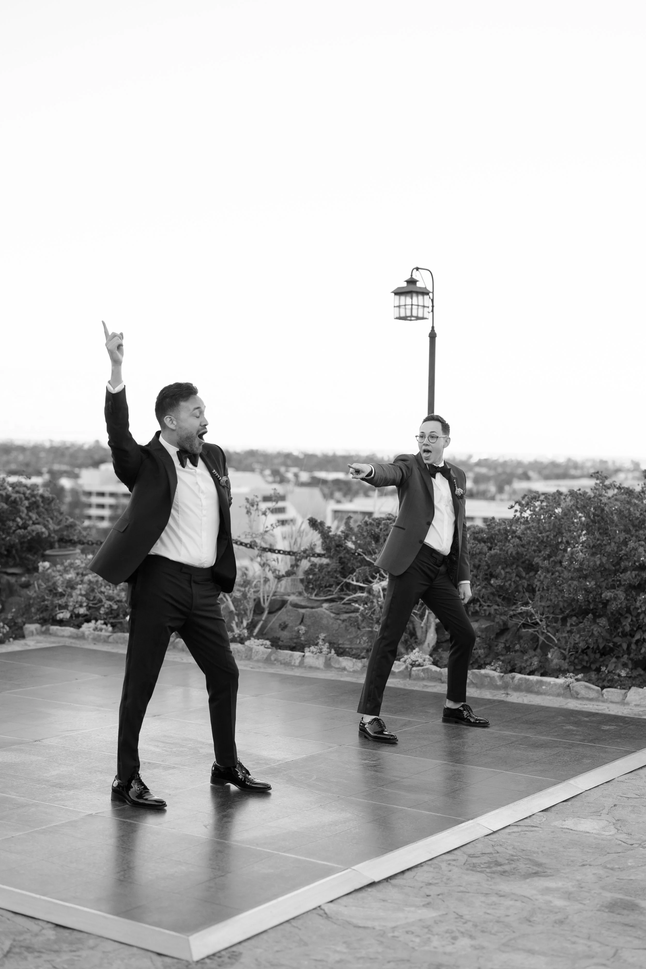 Two men in tuxedos dancing and singing on a small outdoor stage, with one man pointing upwards and the other pointing forward, outdoors with cityscape in the background.