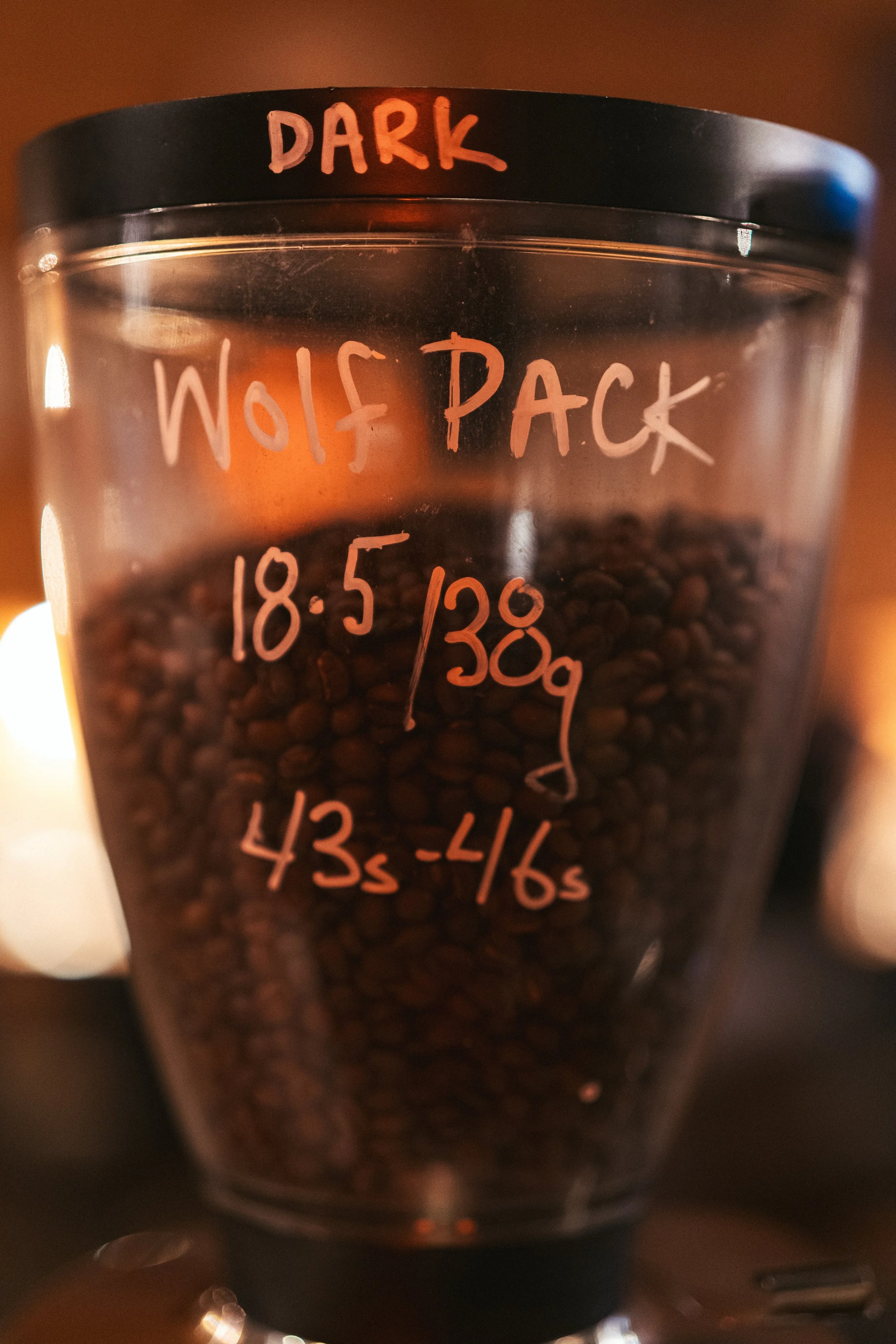 A clear plastic container filled with dark roasted coffee beans, labeled with handwritten text indicating the roast type 'Dark Wolf Pack' and the date '18-5/38g', along with suggested brewing times of 43-46 seconds.