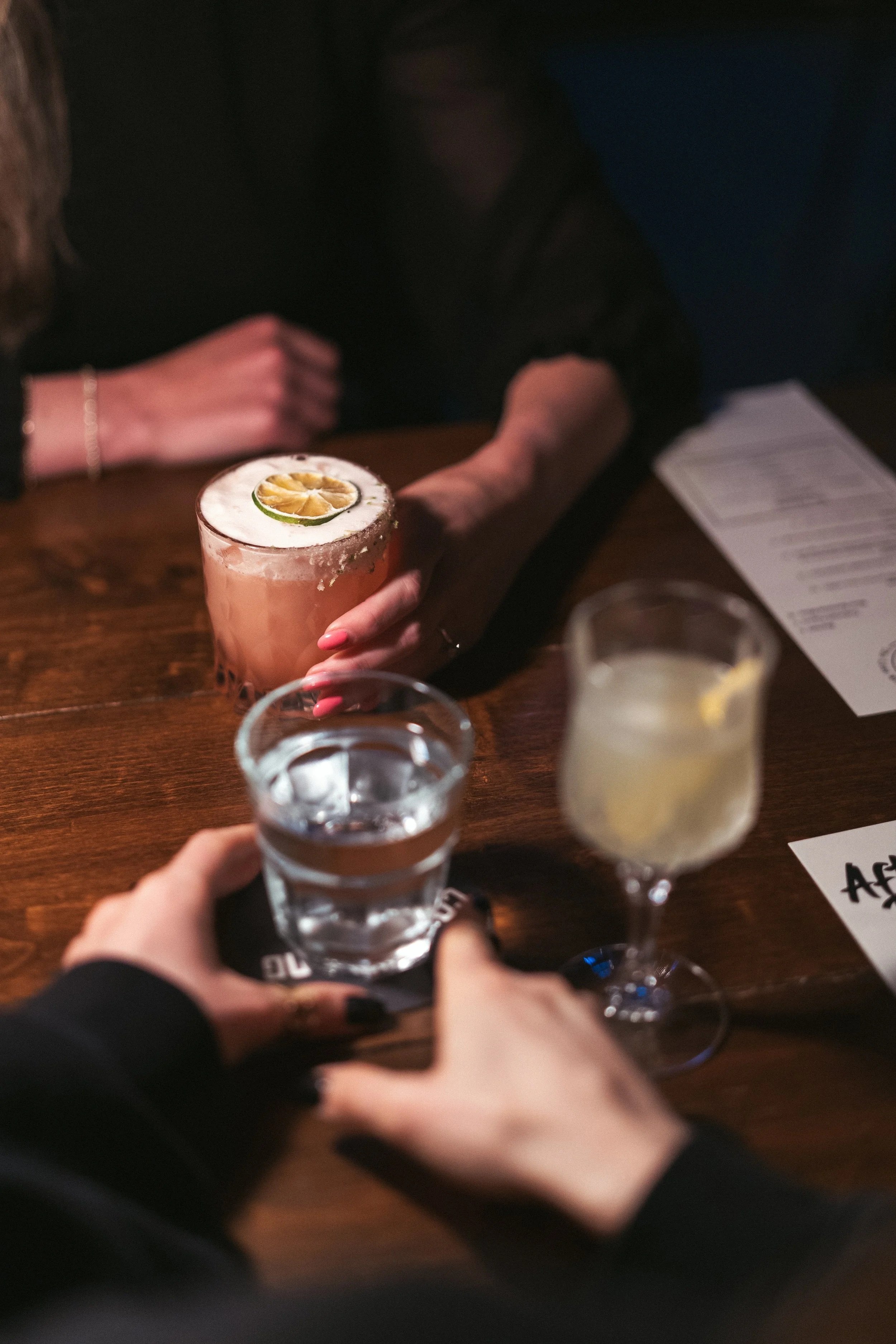 People toasting with drinks at a bar, one person holding a cocktail garnished with a slice of lemon and a lime, another with a glass of water, and a third with a light-colored cocktail.