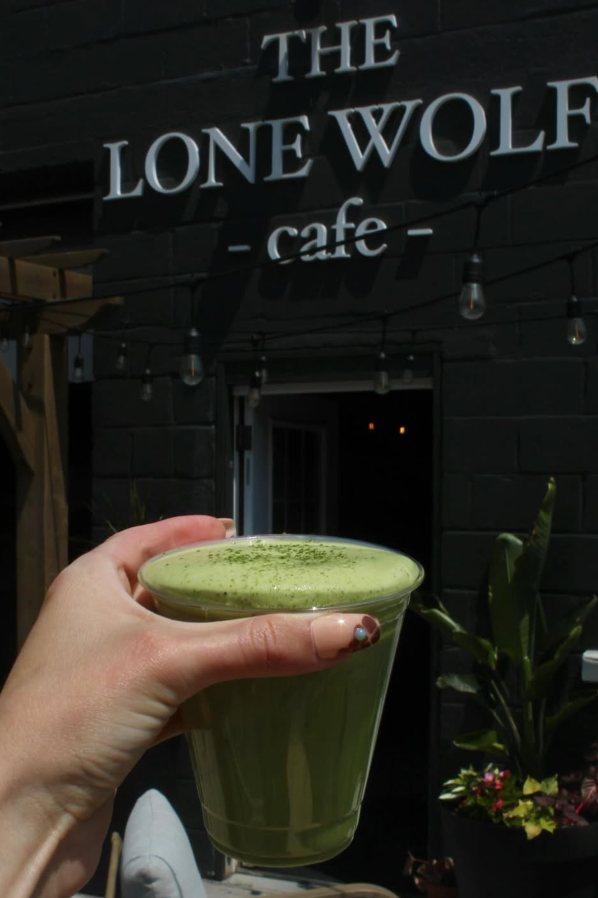A person holding a glass of green matcha latte outside a cafe called 'The Lone Wolf Cafe' at night, with plants visible in the background.
