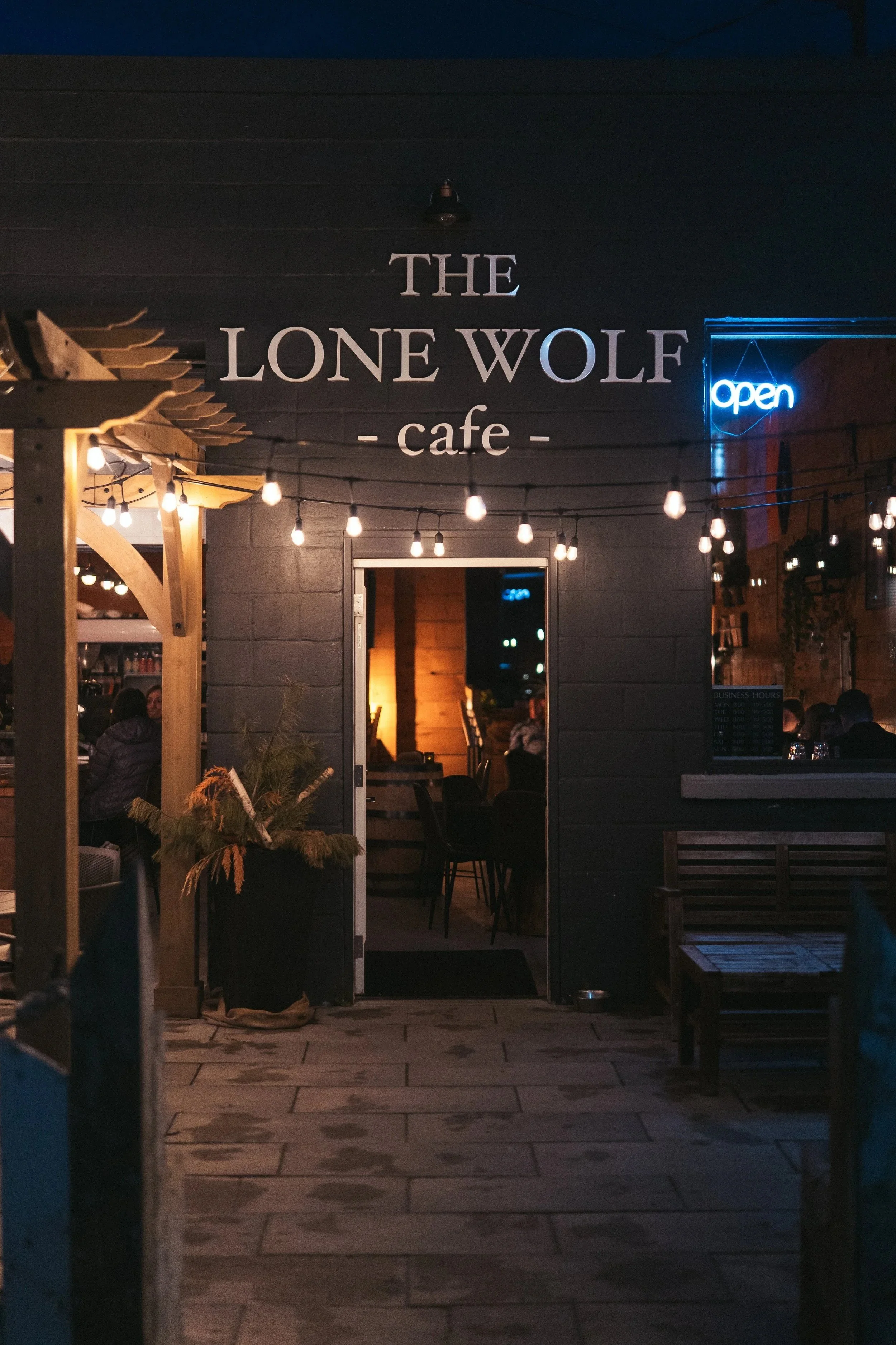 Nighttime scene of the exterior entrance to The Lone Wolf cafe, with string lights hanging above, a neon 'open' sign on the right, and people inside visible through the windows.