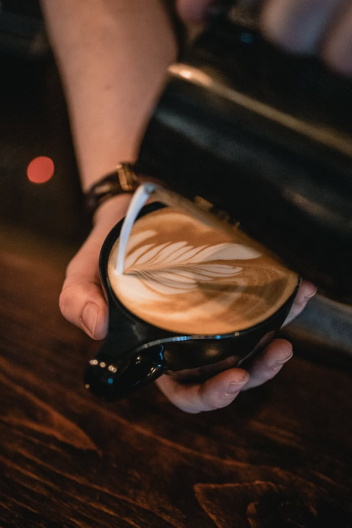 A person pouring steamed milk into a cup of espresso, creating latte art with a leaf design.