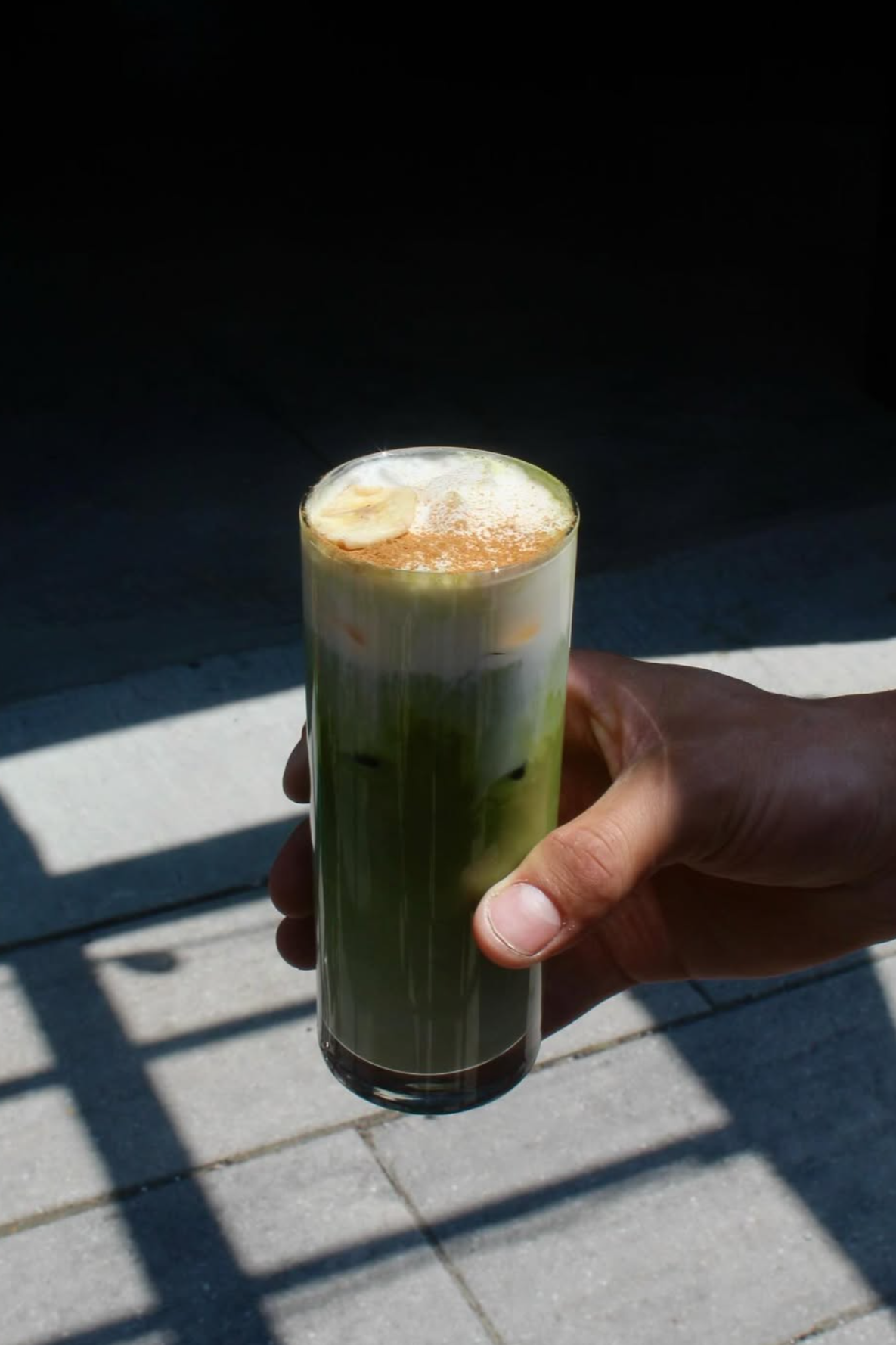 A hand holding a tall glass of layered iced matcha latte with banana slices and foam on top, outdoors on a sunny day.
