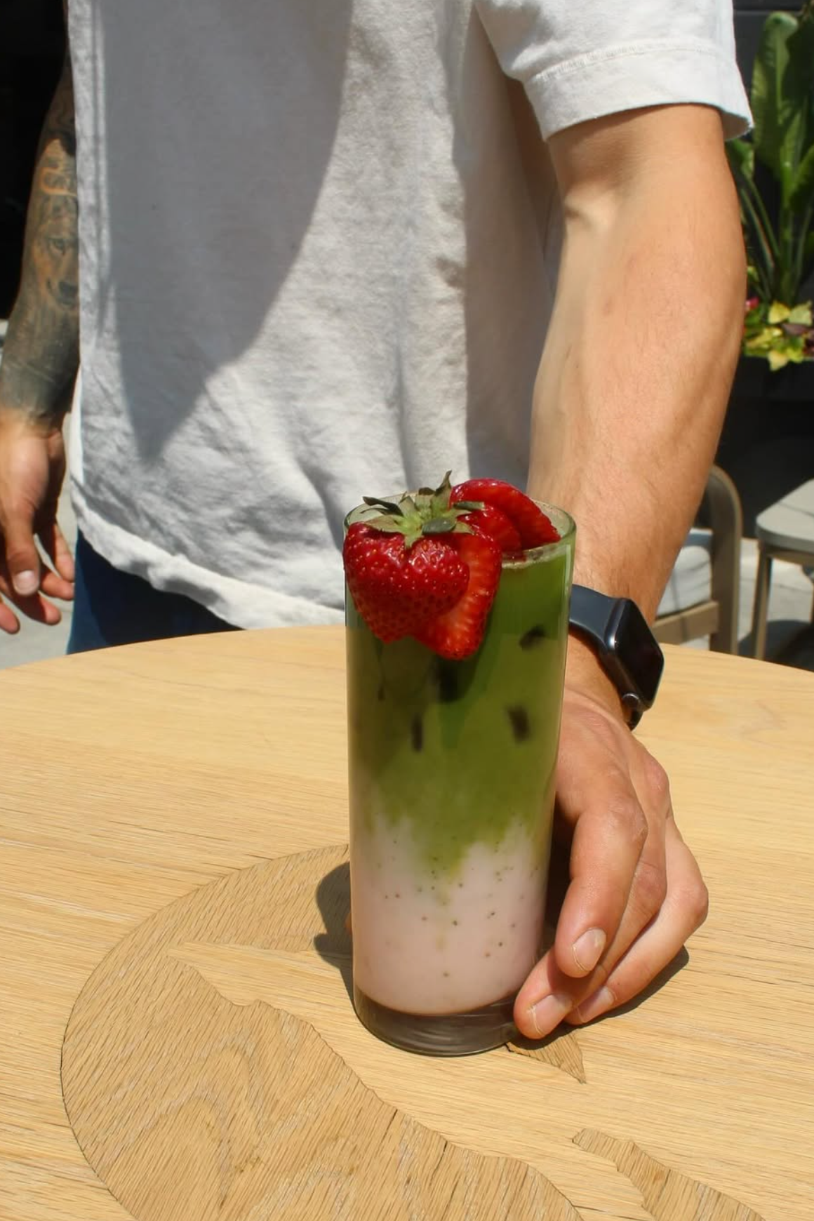 A person holding a tall glass of layered green and white beverage topped with strawberries, sitting at a wooden table outside.