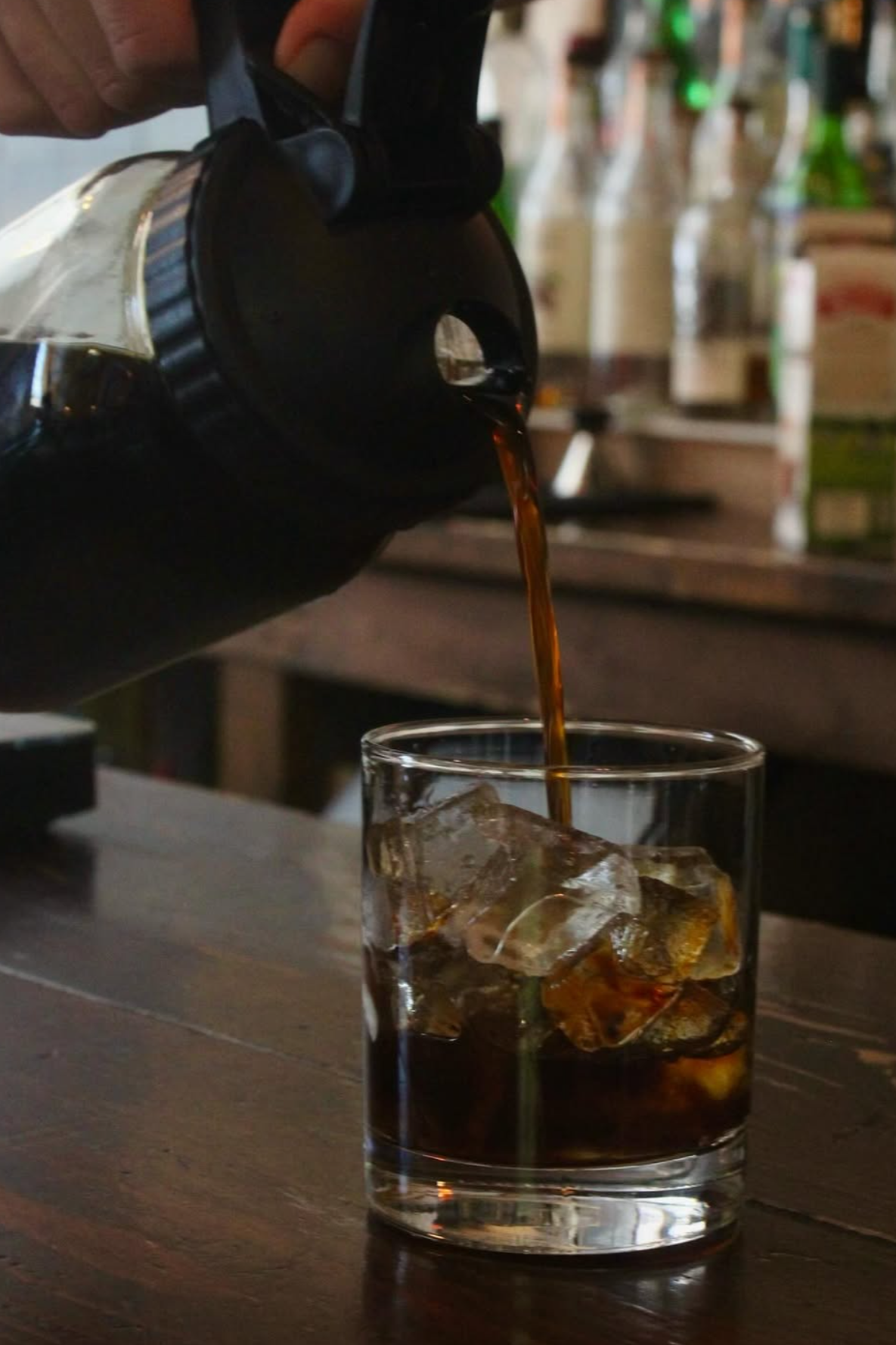 A close-up of a hand pouring cola from a bottle into a glass filled with ice on a wooden surface.