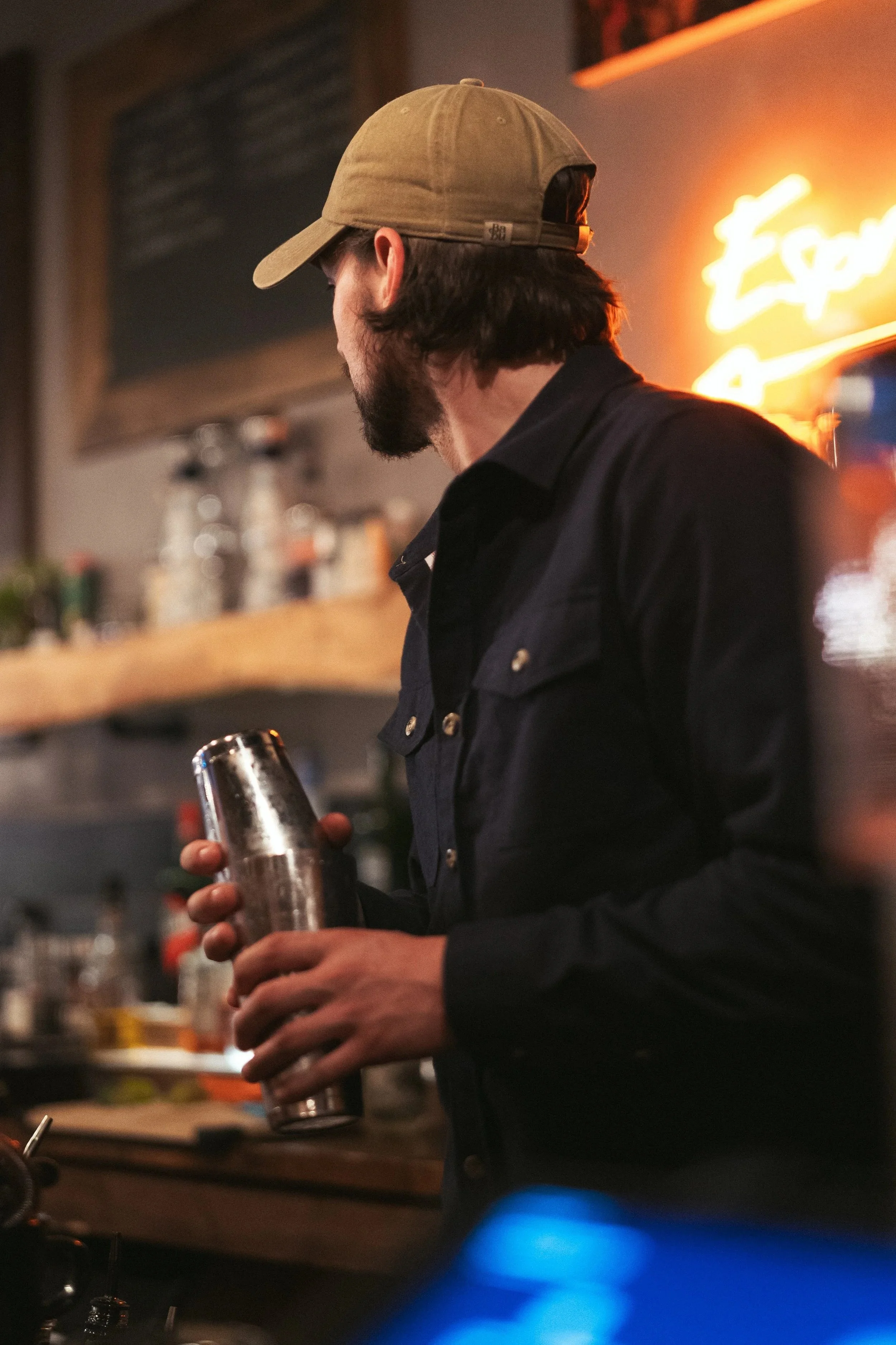A man with long dark hair, beard, wearing a tan baseball cap and a dark shirt, holding a metal shaker in a dimly lit bar or restaurant.